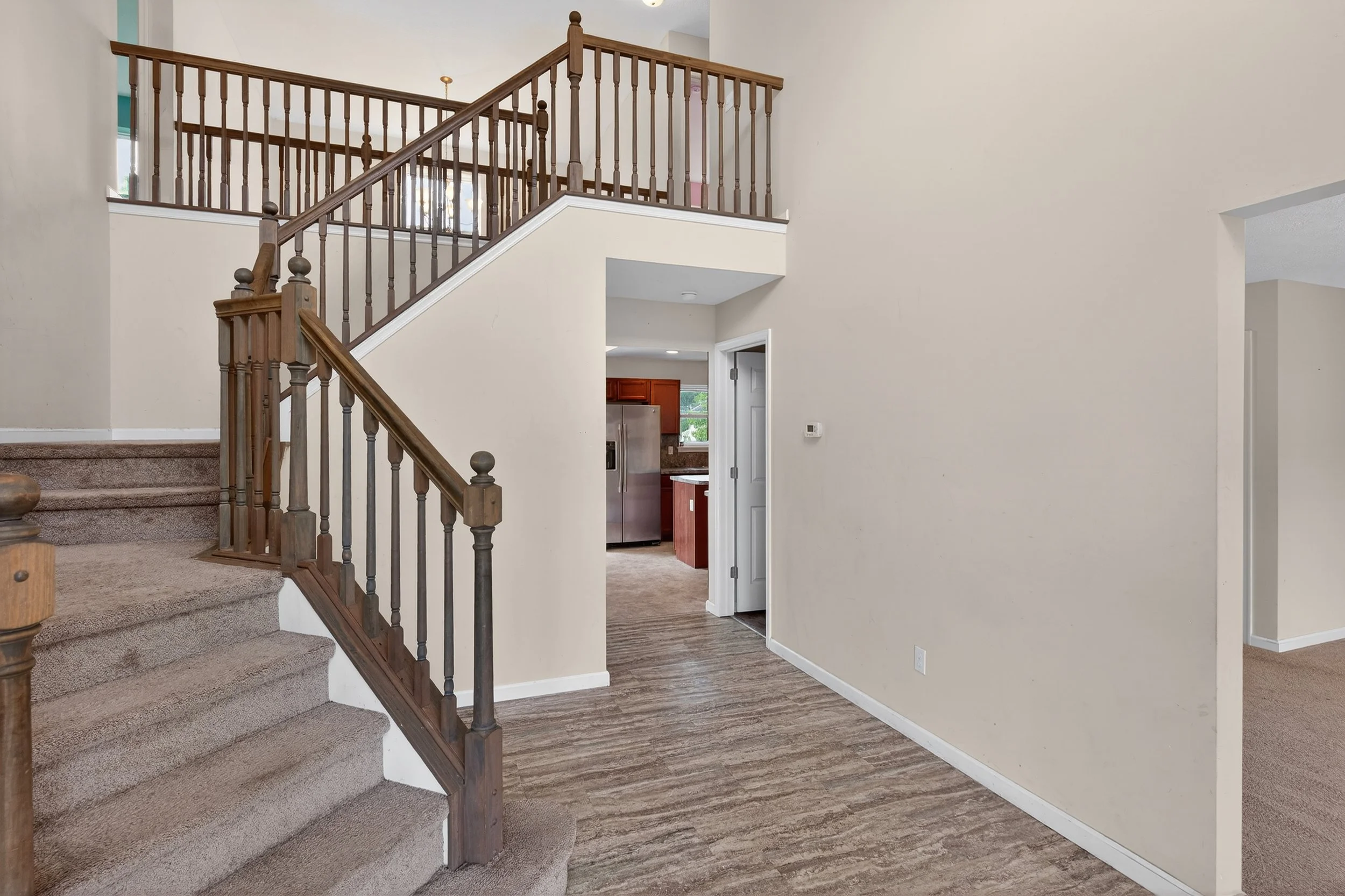 Interior view of a house showing a staircase with wooden railing, open entry to a kitchen with wooden cabinets and a stainless steel refrigerator, and beige walls with a thermostat on the wall.