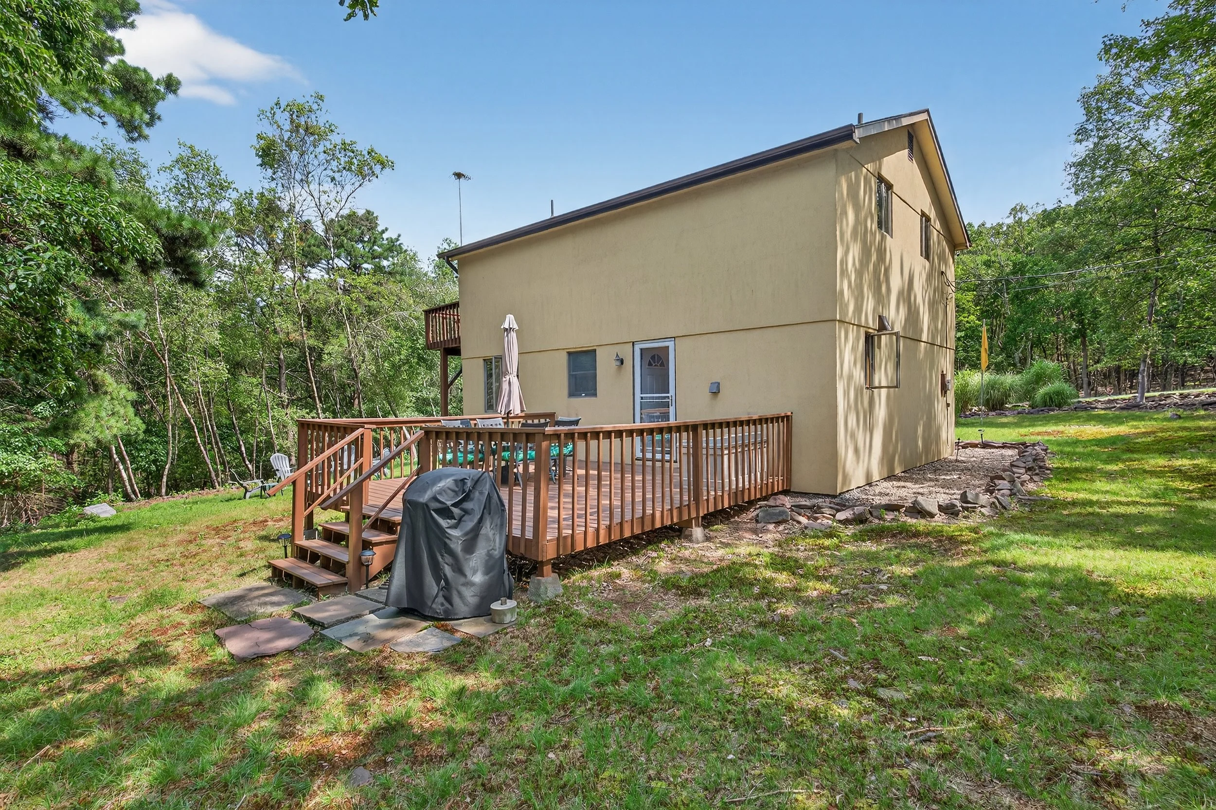 Backyard view of a two-story yellow house with a wooden deck, outdoor furniture, a grill covered with a protective cover, and surrounded by green trees and grass.