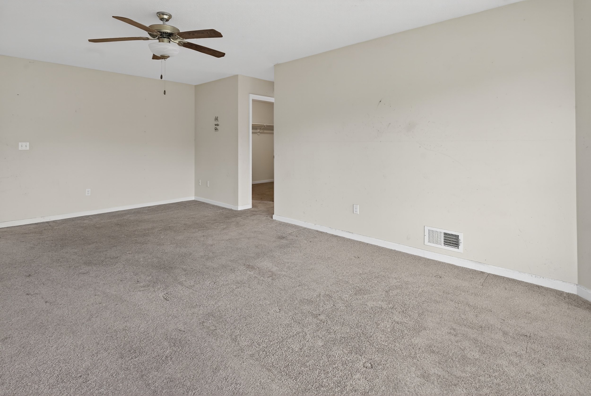 Empty living room with beige carpet, off-white walls, ceiling fan, and an open door leading to a closet.