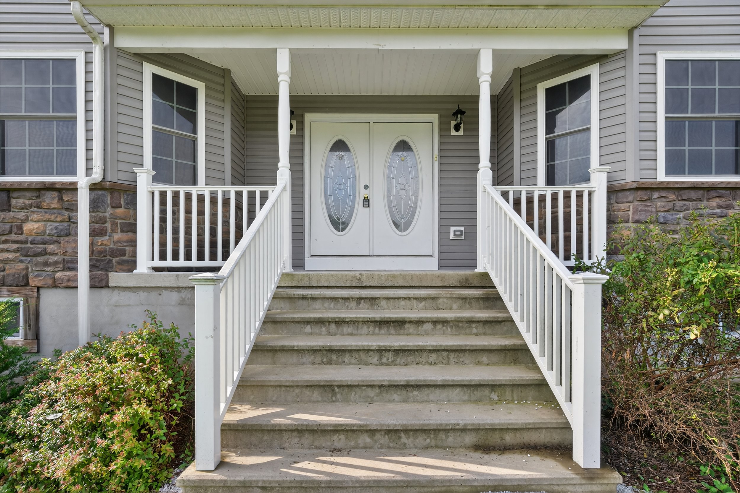 Front door of a house with stairs, white double door with oval glass panels, gray vinyl siding, stone accents, and two windows, surrounded by bushes.