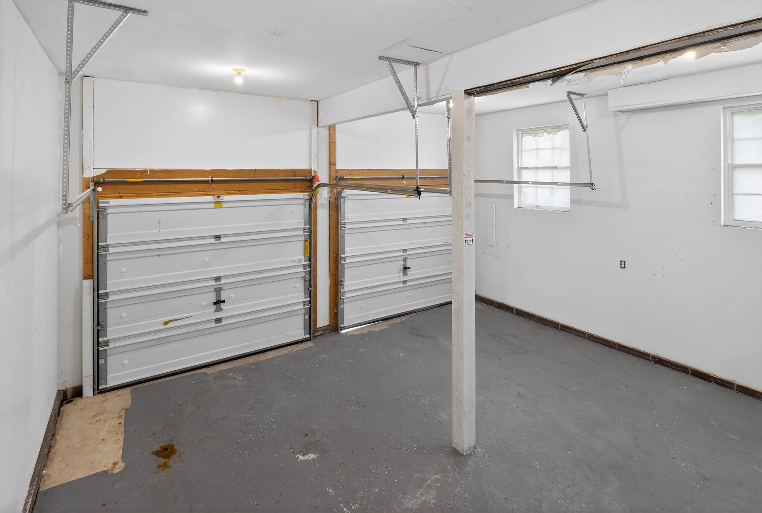 Empty garage with white double doors, unpainted concrete floor, and two small windows, with one ceiling light and a support column in the center.