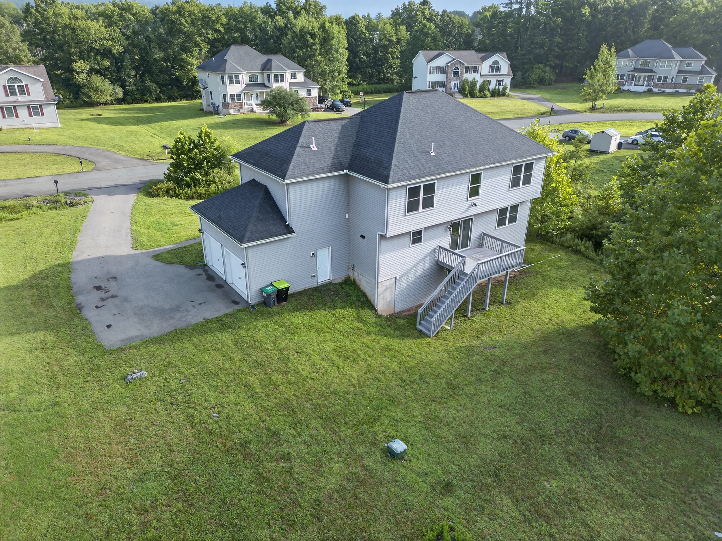 An aerial view of a two-story house with a deck and stairs at the back, white siding, gray roof, and a driveway with two trash bins. The house is surrounded by green grass and other similar houses in the background, with trees and a wooded area behin