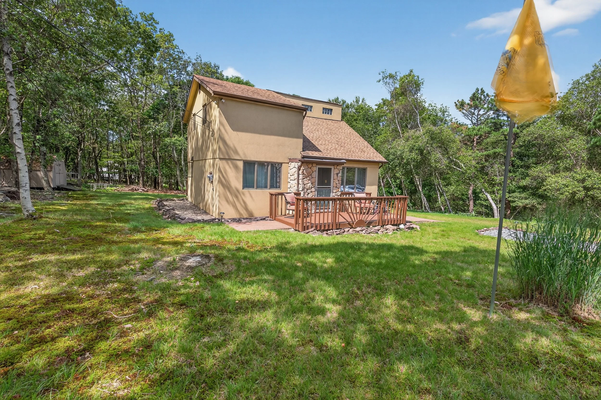 Backyard view of a beige house with a small wooden deck, surrounded by green grass and trees, with a yellow umbrella on the right side.