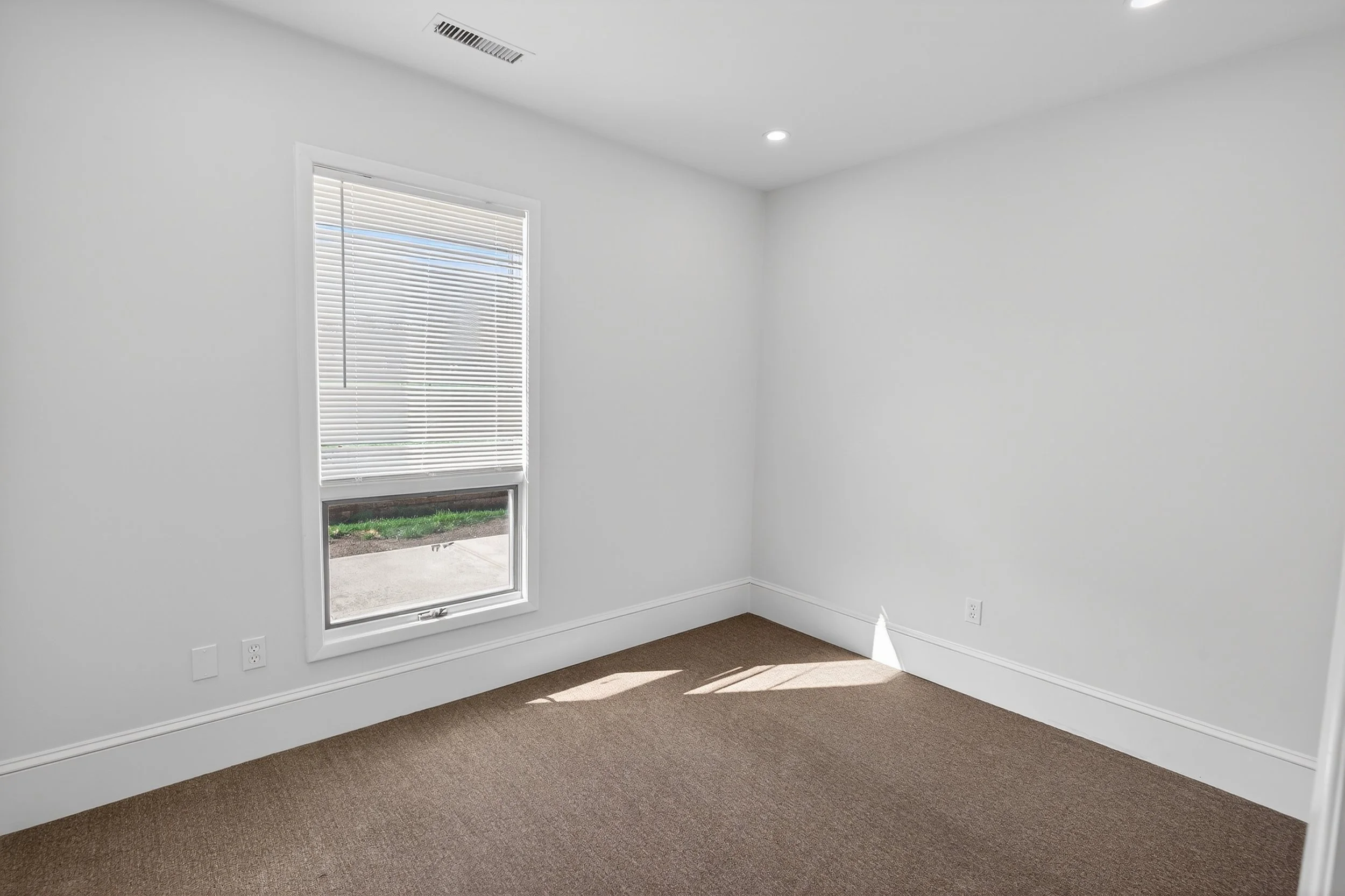 Empty room with white walls, brown carpet, a window with blinds, and a ceiling light