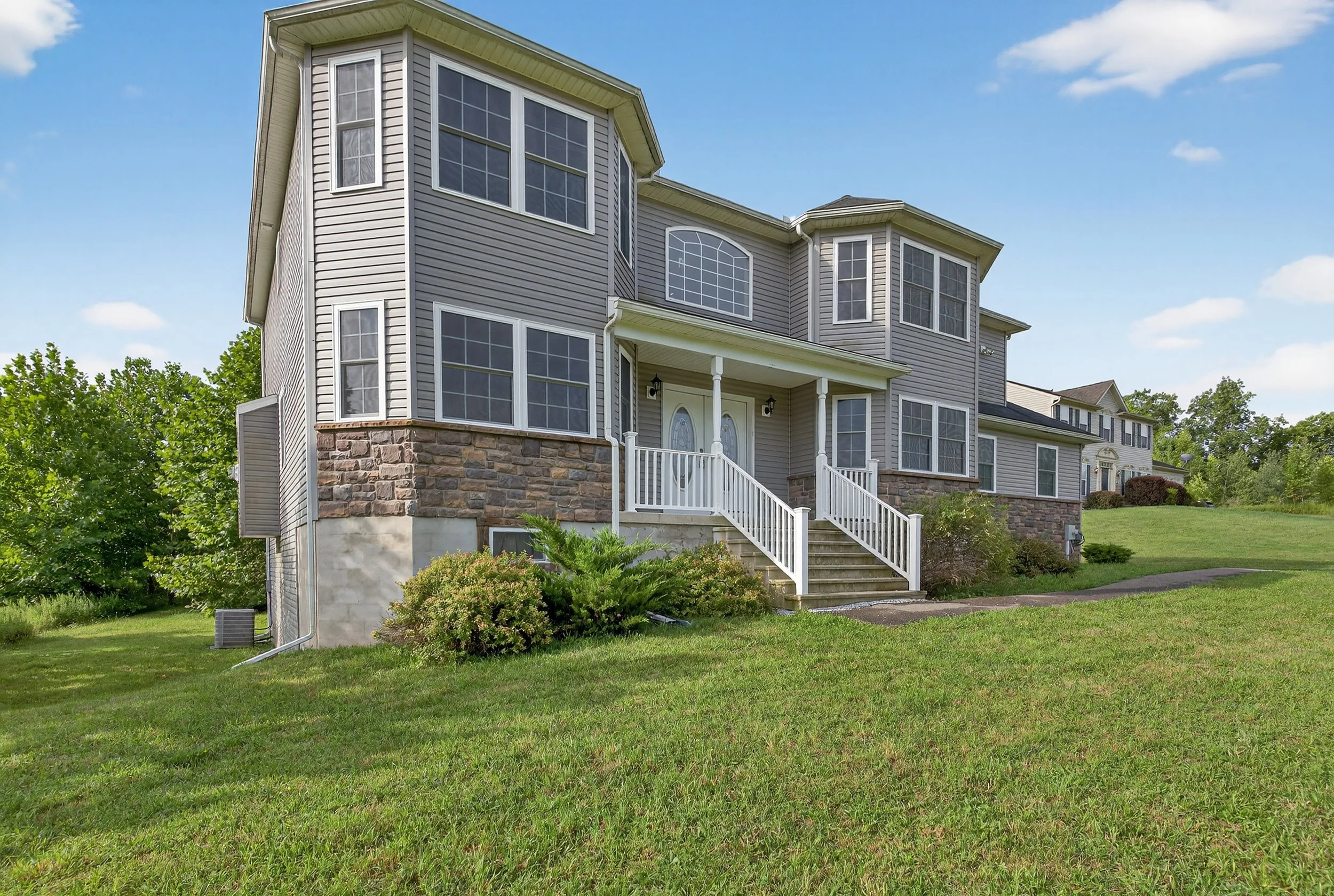 Large two-story house with grey siding and stone accents, white stairs leading to front door, multiple large windows, green lawn, and nearby trees, under a partly cloudy sky.