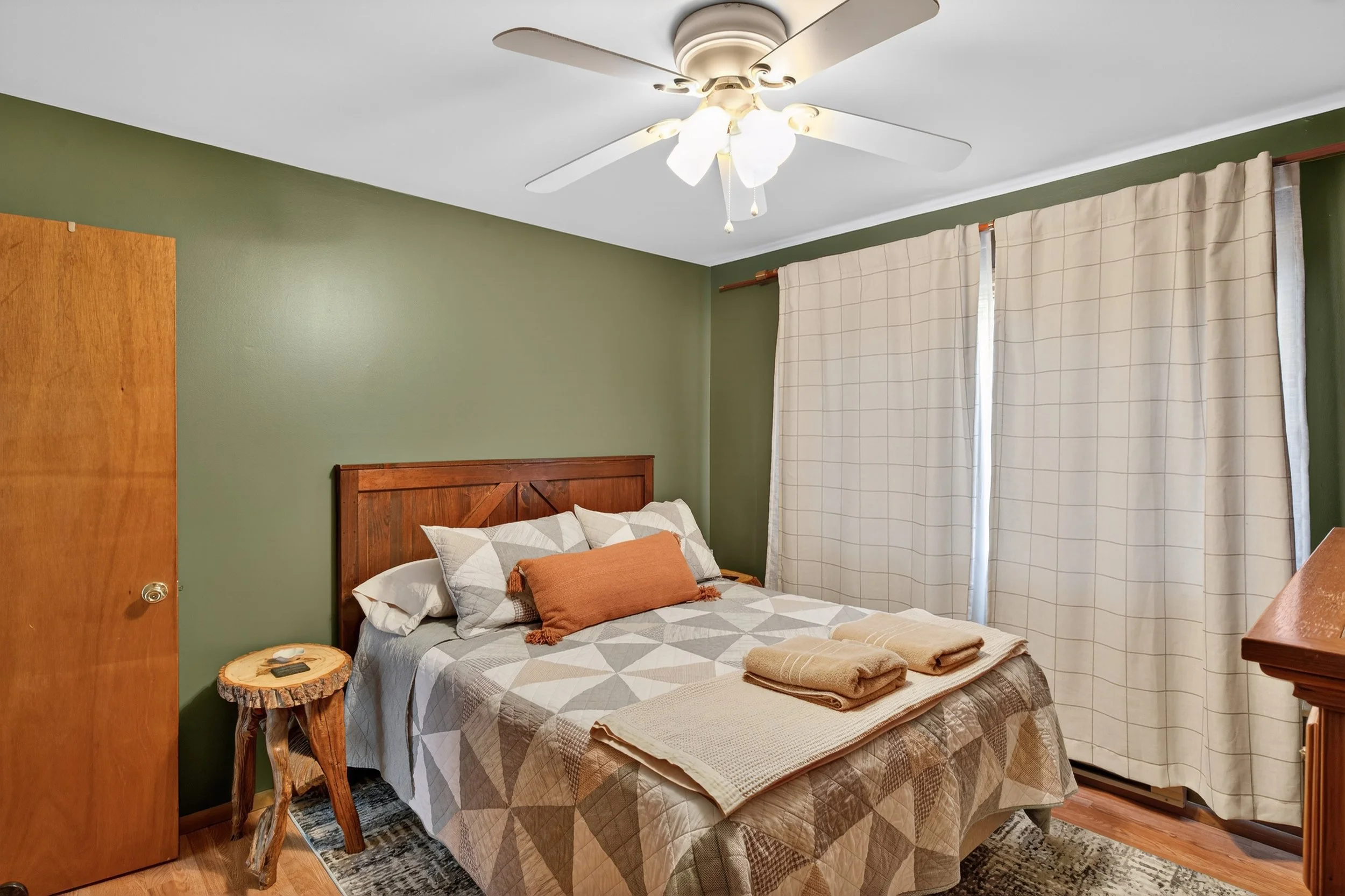 Bedroom with green wall, wooden bed with geometric quilt, two pillows, a rolled orange pillow, towels on bed, wooden side table, cream curtains, ceiling fan, hardwood floor, and area rug.