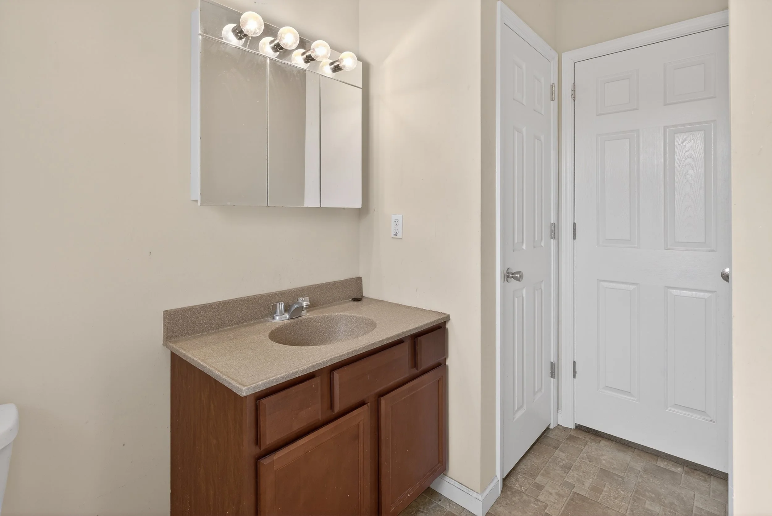 Bathroom with beige walls, a brown wooden vanity with a beige countertop, a mirror cabinet, a white door, and a white closet door.