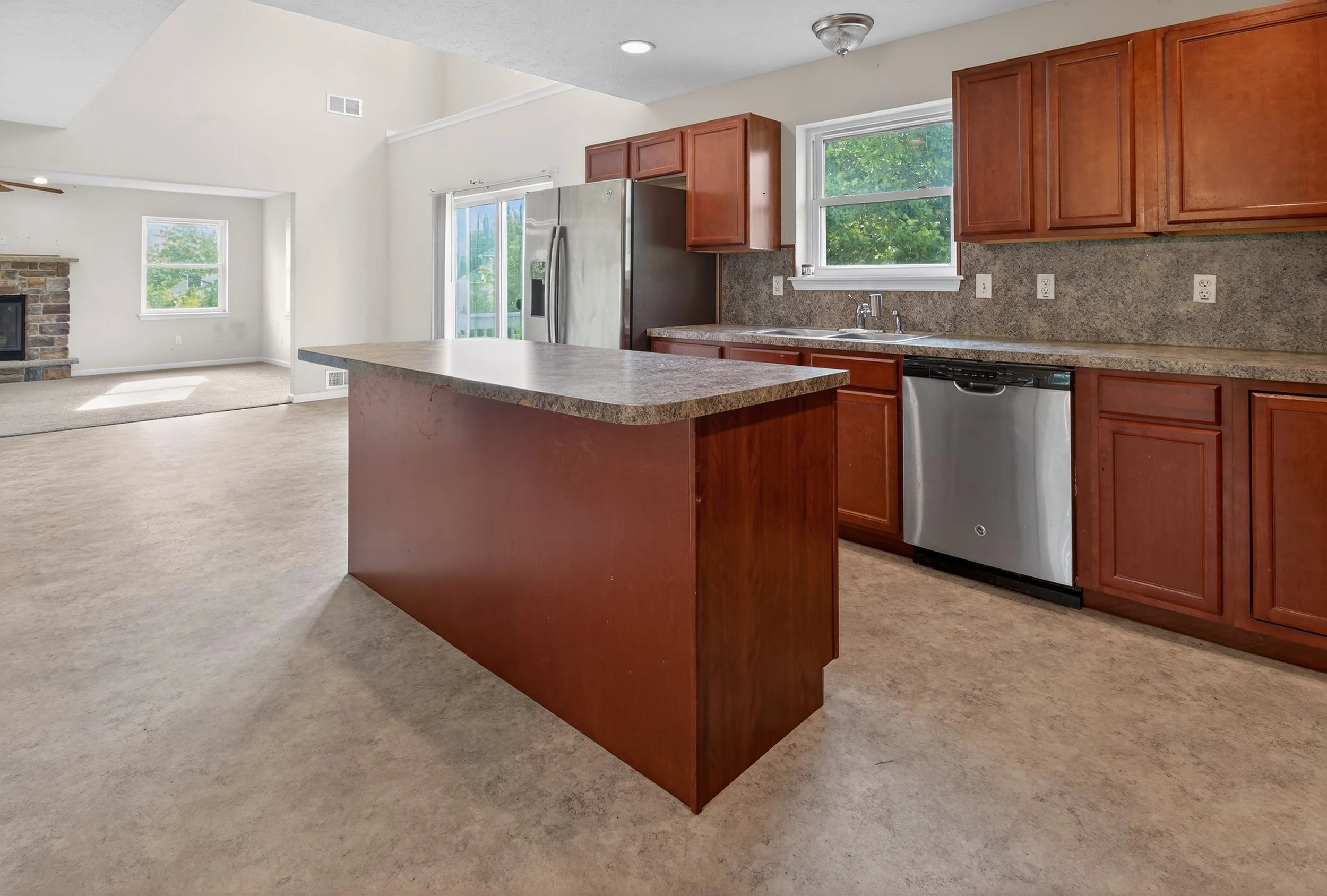 Open-concept kitchen with brown wooden cabinets, granite countertops, stainless steel appliances, and a large island, adjacent to a living area with a fireplace and a sliding glass door leading outside.