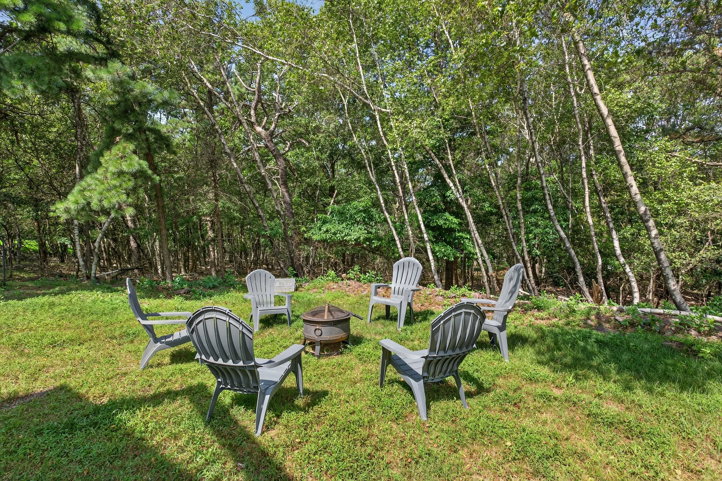 Five gray Adirondack chairs arranged in a circle around a metal fire pit on a grassy yard, with a dense green forest in the background.