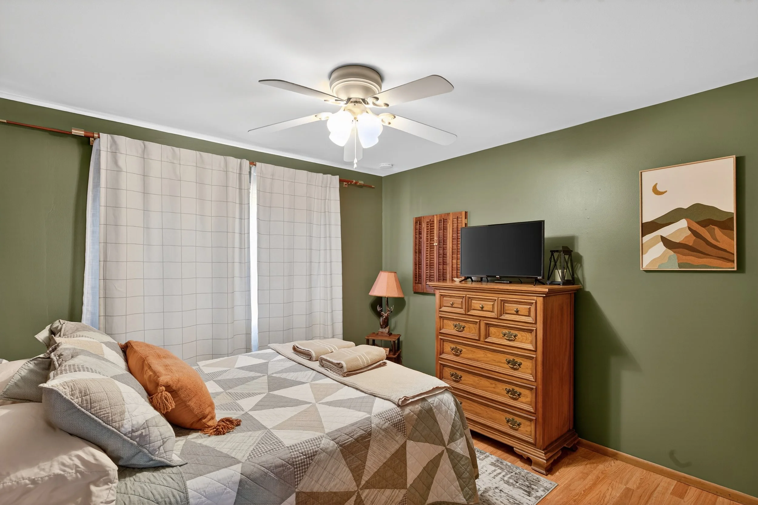 A cozy bedroom with a green wall, a ceiling fan, a bed with patterned bedding and pillows, a dresser with a TV, a bedside table with a lamp, curtains covering the window, and a framed landscape painting.