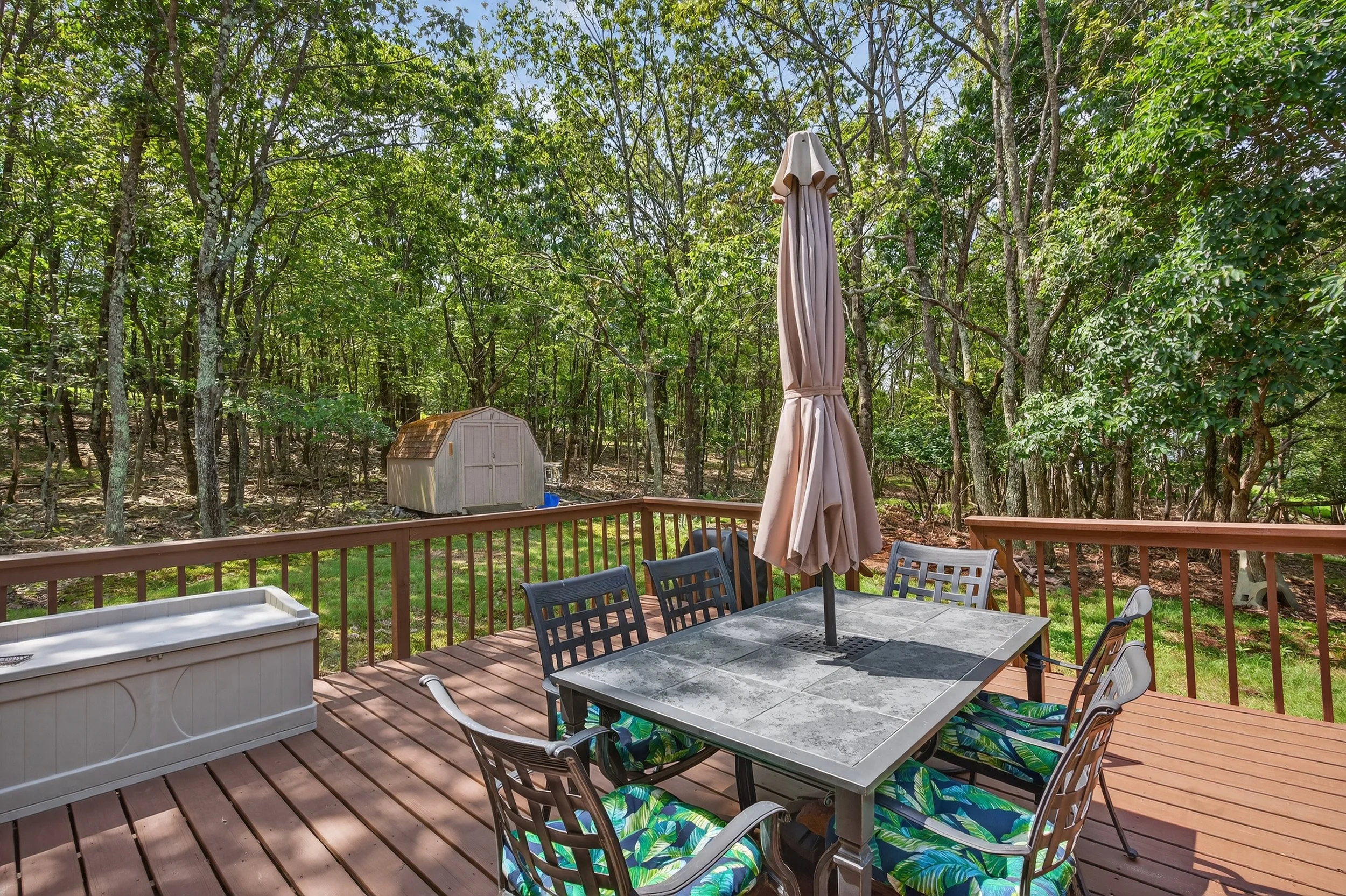 A wooden deck with a table, five chairs with patterned cushions, a large closed umbrella, and a privacy fence, overlooking a backyard with trees and a small shed.