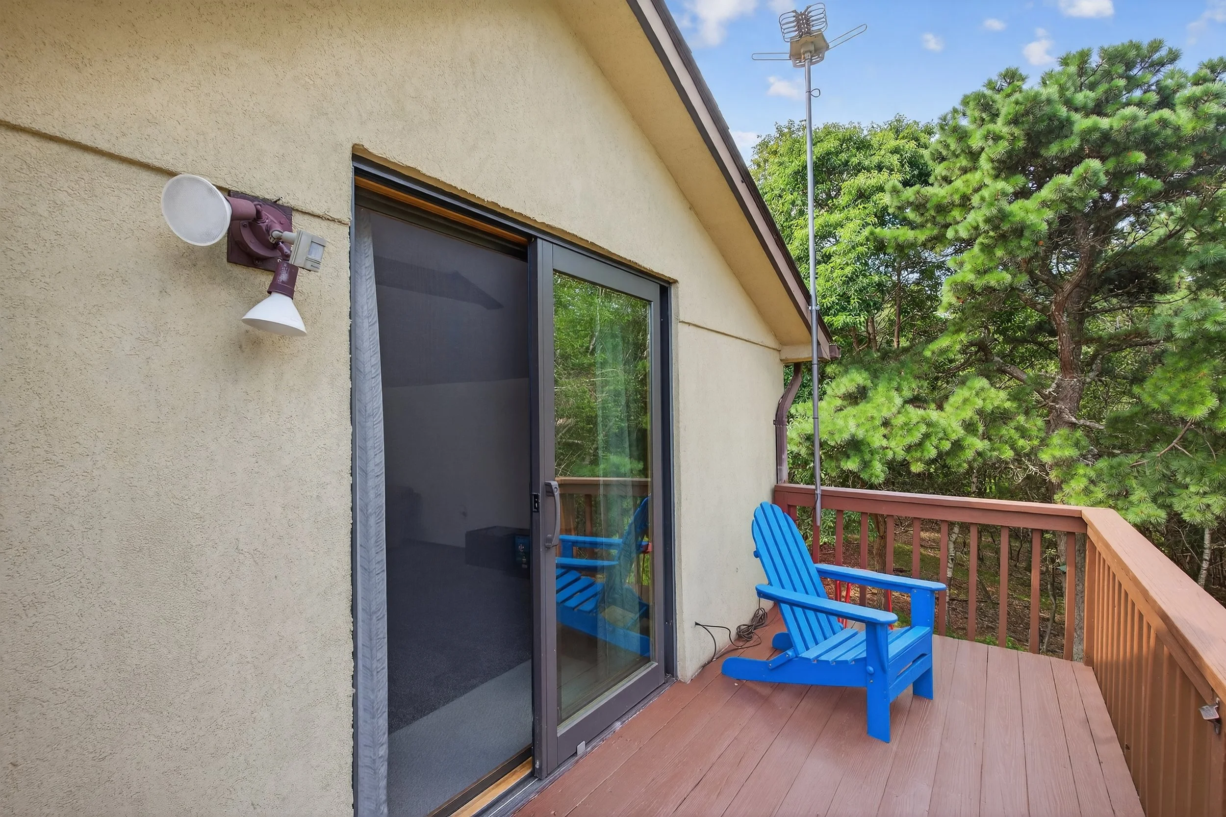 A small outdoor balcony with a blue Adirondack chair on a wooden deck, beige building wall with an outdoor light fixture, sliding glass door, and a TV antenna on the roof surrounded by green trees and a blue sky.