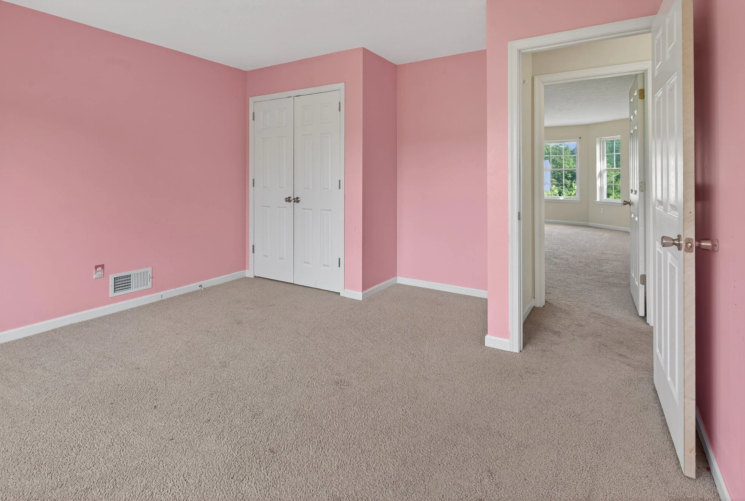 Empty pink-walled bedroom with beige carpet, white double closet doors, and an open doorway leading to another room with beige walls, carpet, and windows.