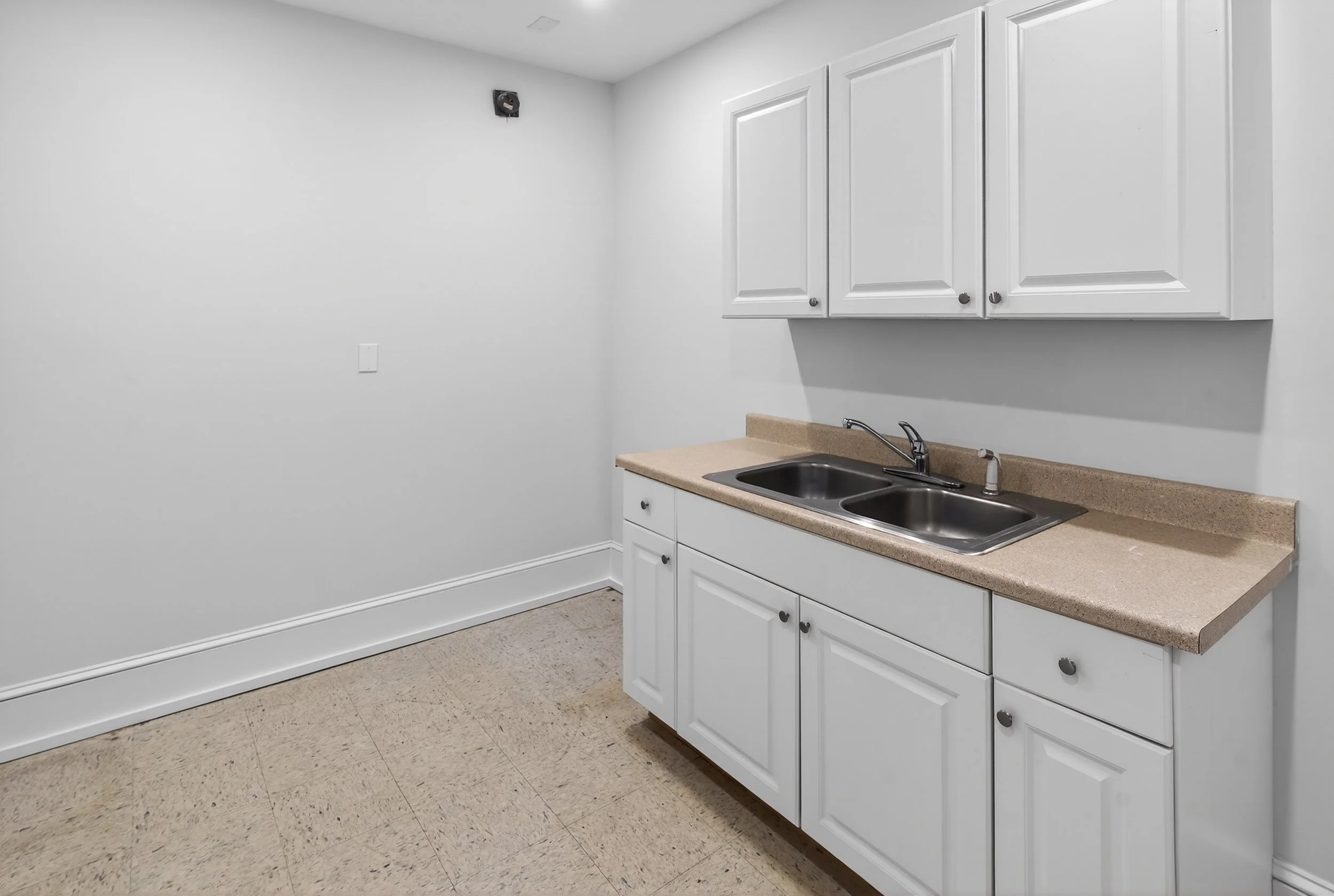 A small kitchen area with white cabinets, a beige countertop, and a double stainless steel sink. The walls are plain white with an electrical outlet and a small black device mounted near the ceiling, and the floor is tan with a slightly textured surf