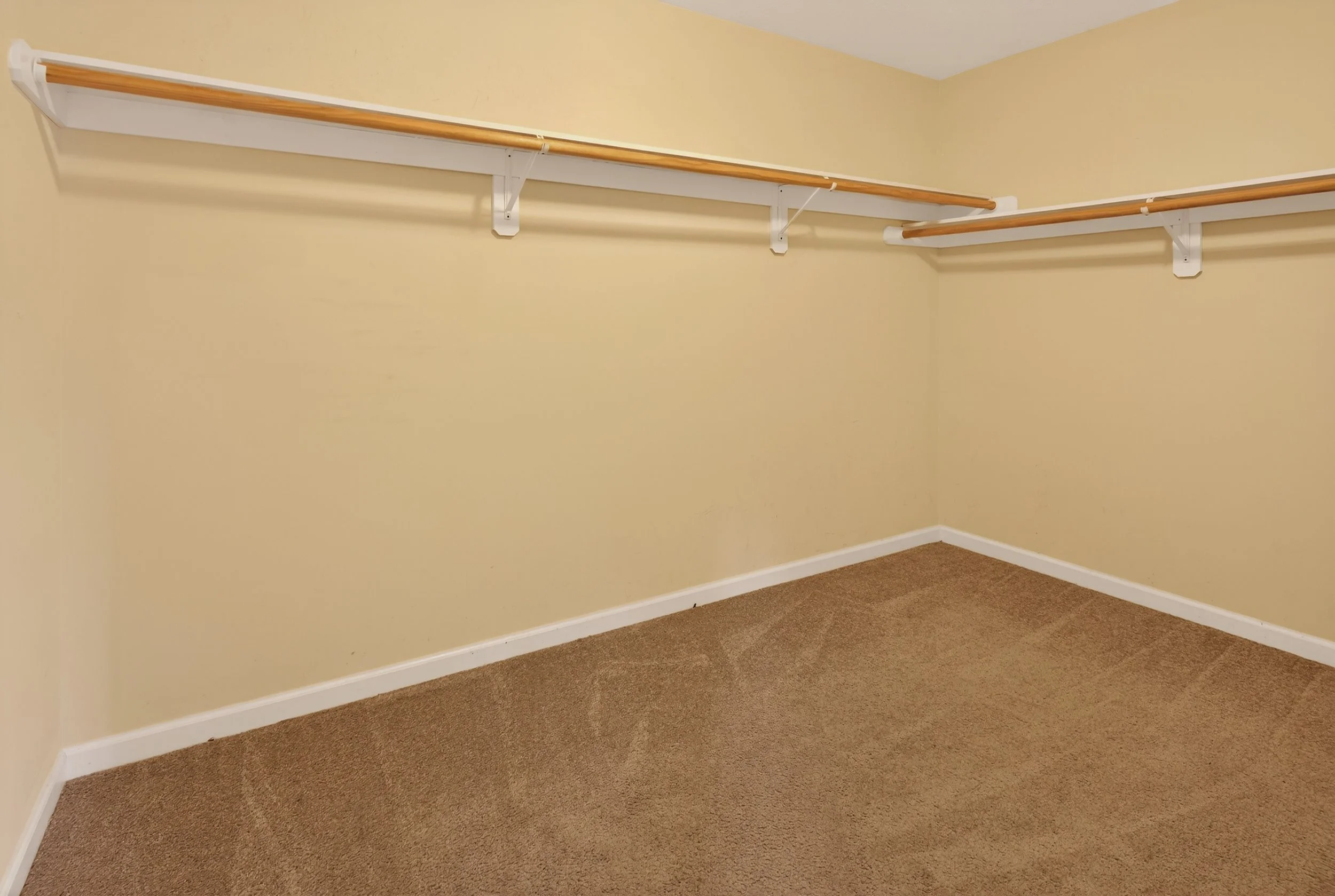 Empty walk-in closet with beige walls, carpeted floor, and two wooden clothing rods mounted on white brackets.