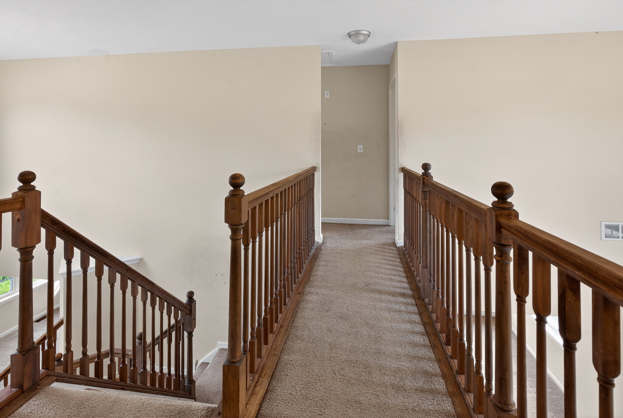 Interior view of a second-floor hallway with wooden railings and beige walls, leading to a small doorway at the end of the corridor.