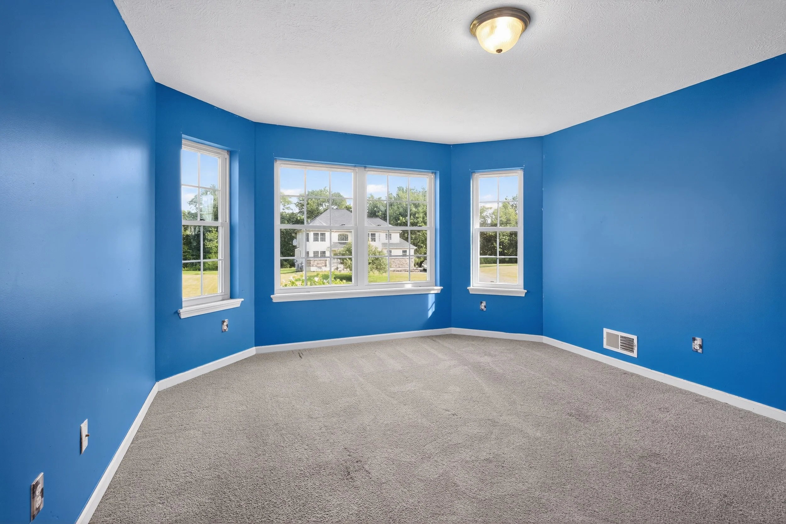 Empty room with blue walls, three large windows, beige carpet, and white trim, with a ceiling light fixture.