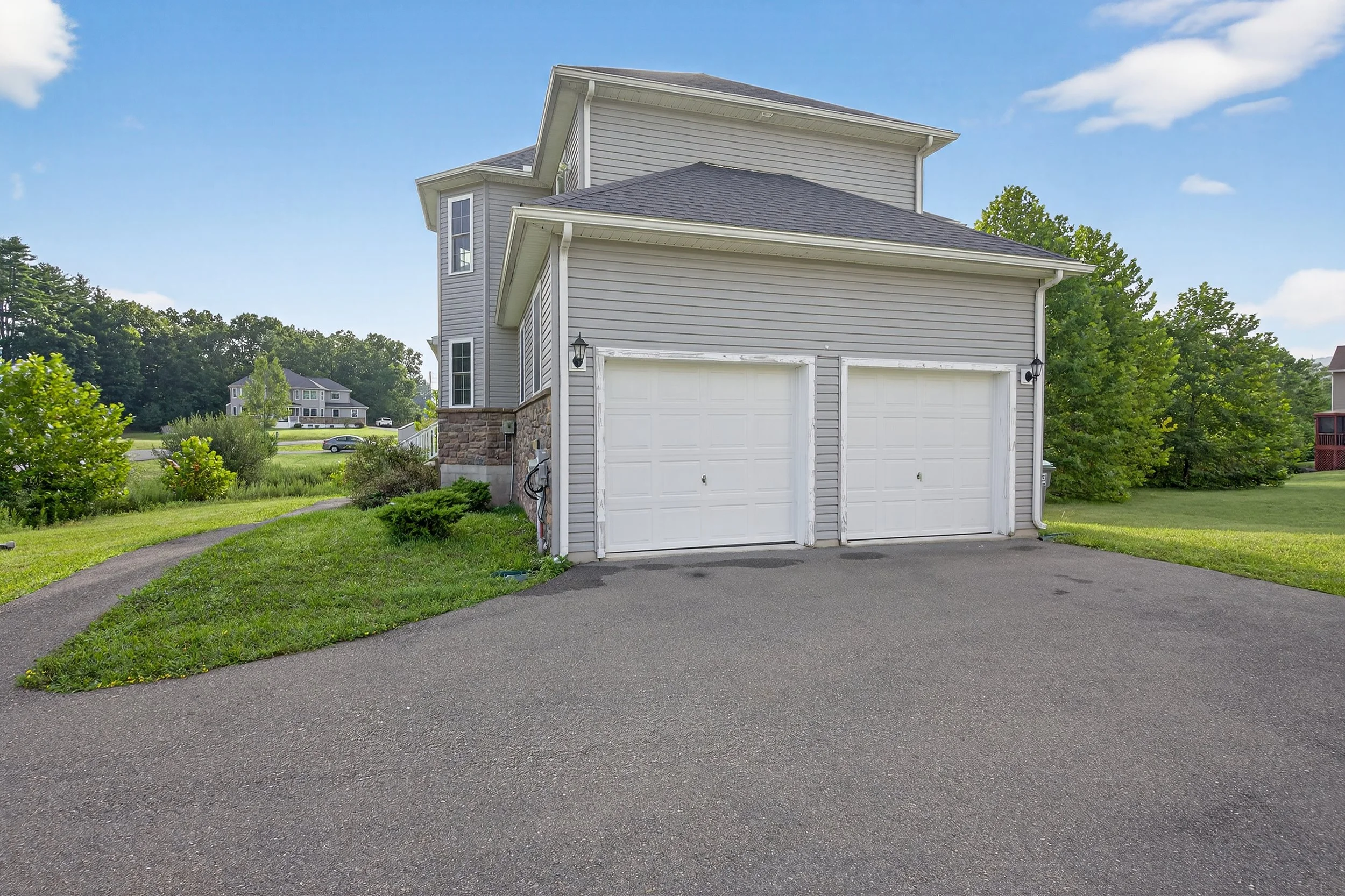 A two-story house with a garage, beige siding, and a stone foundation, surrounded by green lawn and trees under a blue sky.