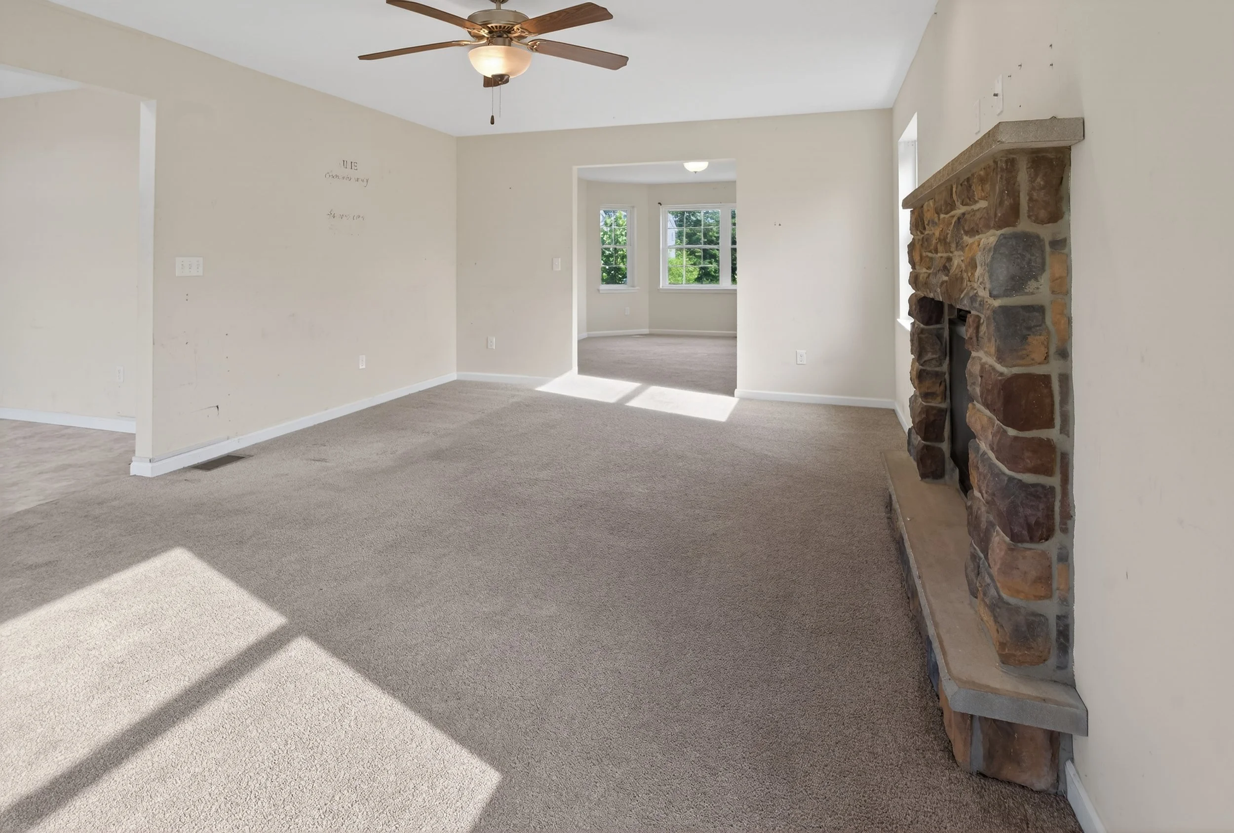 Living room with beige walls, carpeted floor, stone fireplace, ceiling fan, and large windows.