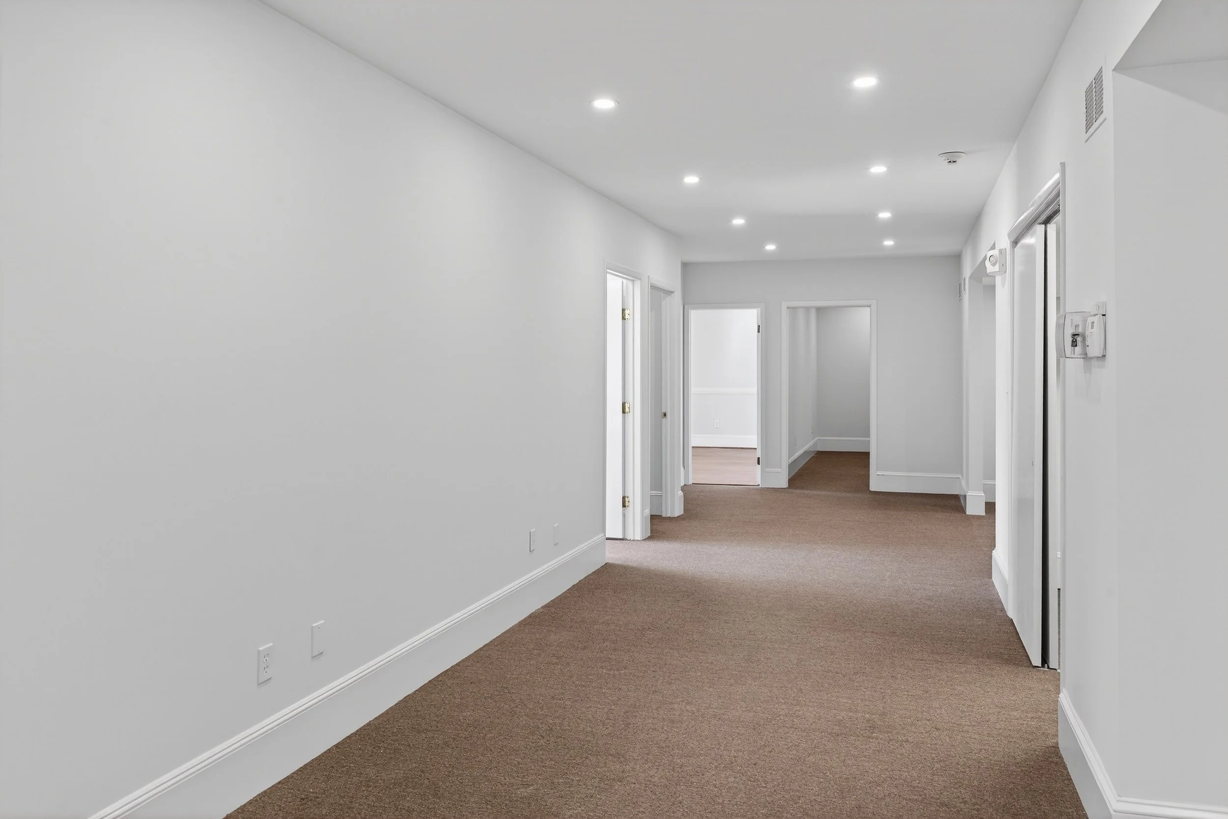 Empty white-walled corridor with brown carpet, multiple doorways, and ceiling lights.