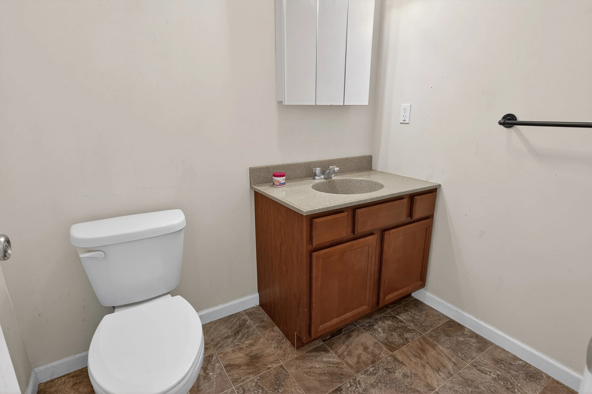 Small bathroom with white toilet, wooden vanity with beige countertop and sink, mirror cabinet, black towel bar, and tiled floor.