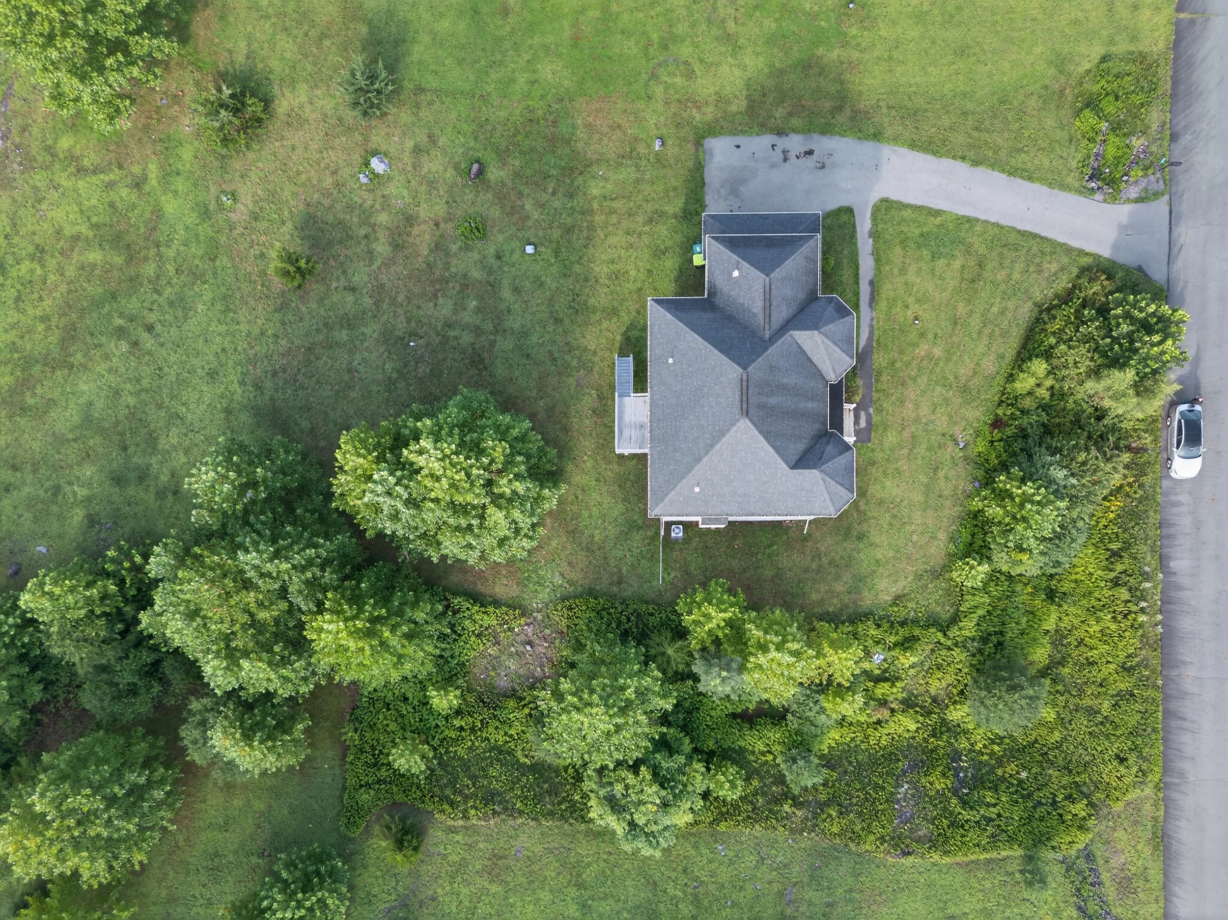 Top-down aerial view of a gray house with a porch, situated on a grassy yard with trees and bushes around, and a driveway leading to a street with a parked white vehicle.