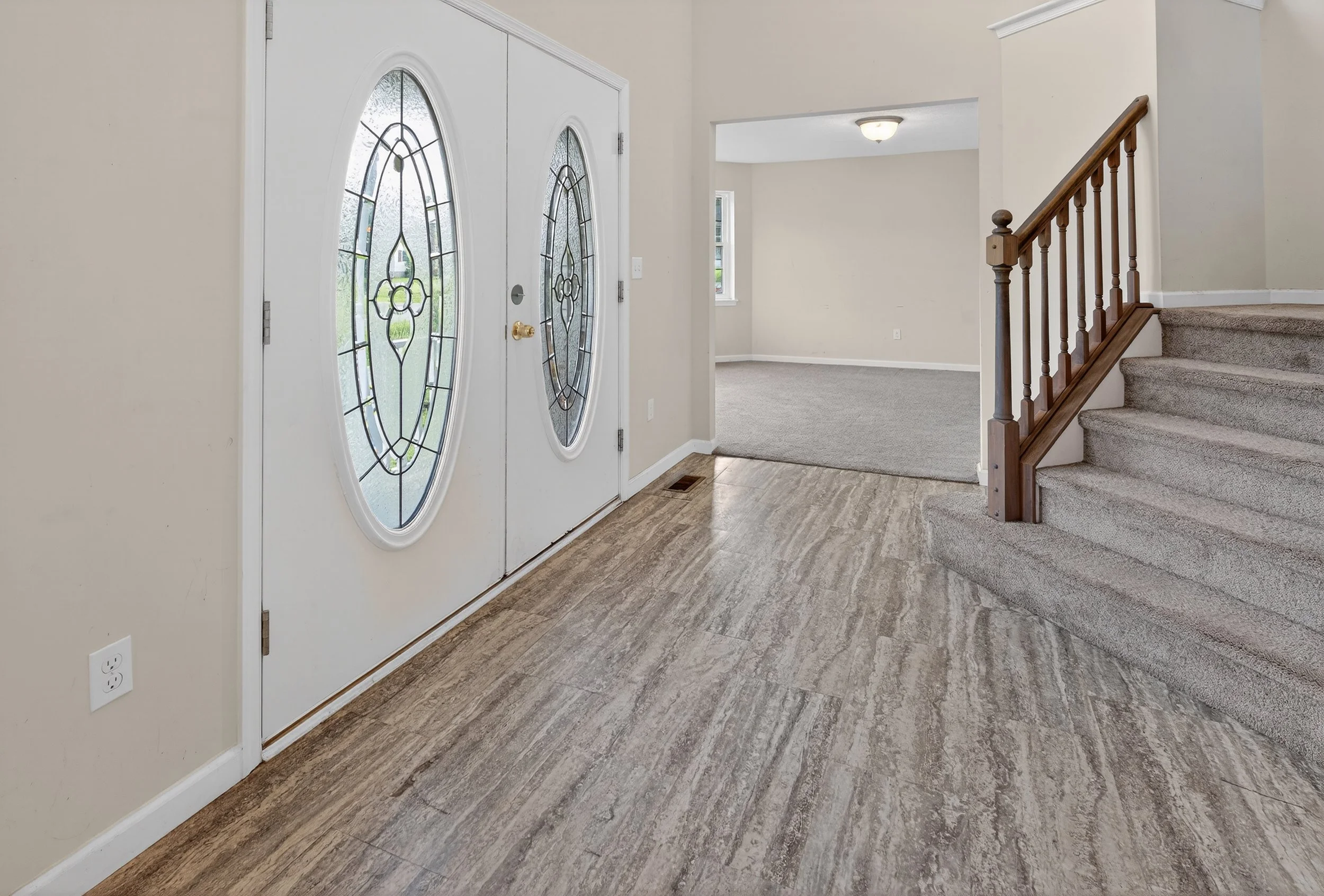 Entryway with white front door featuring oval glass panes and a brass doorknob, leading to a carpeted living room with a staircase and wooden handrail, beige walls, gray flooring, and natural light through a window.