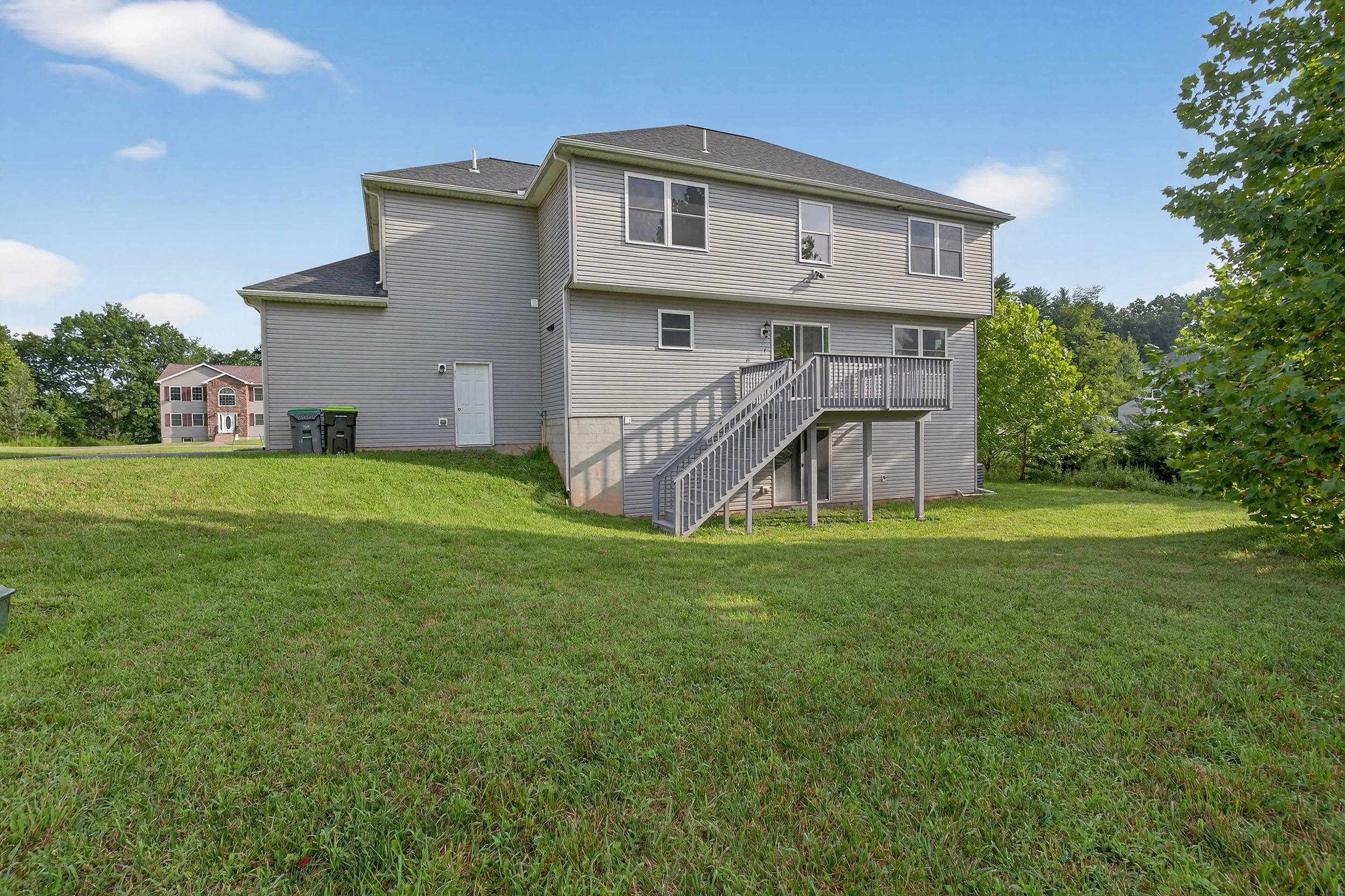 Back of a two-story house with gray siding, a deck with stairs, windows, and a lawn with green grass and trees.