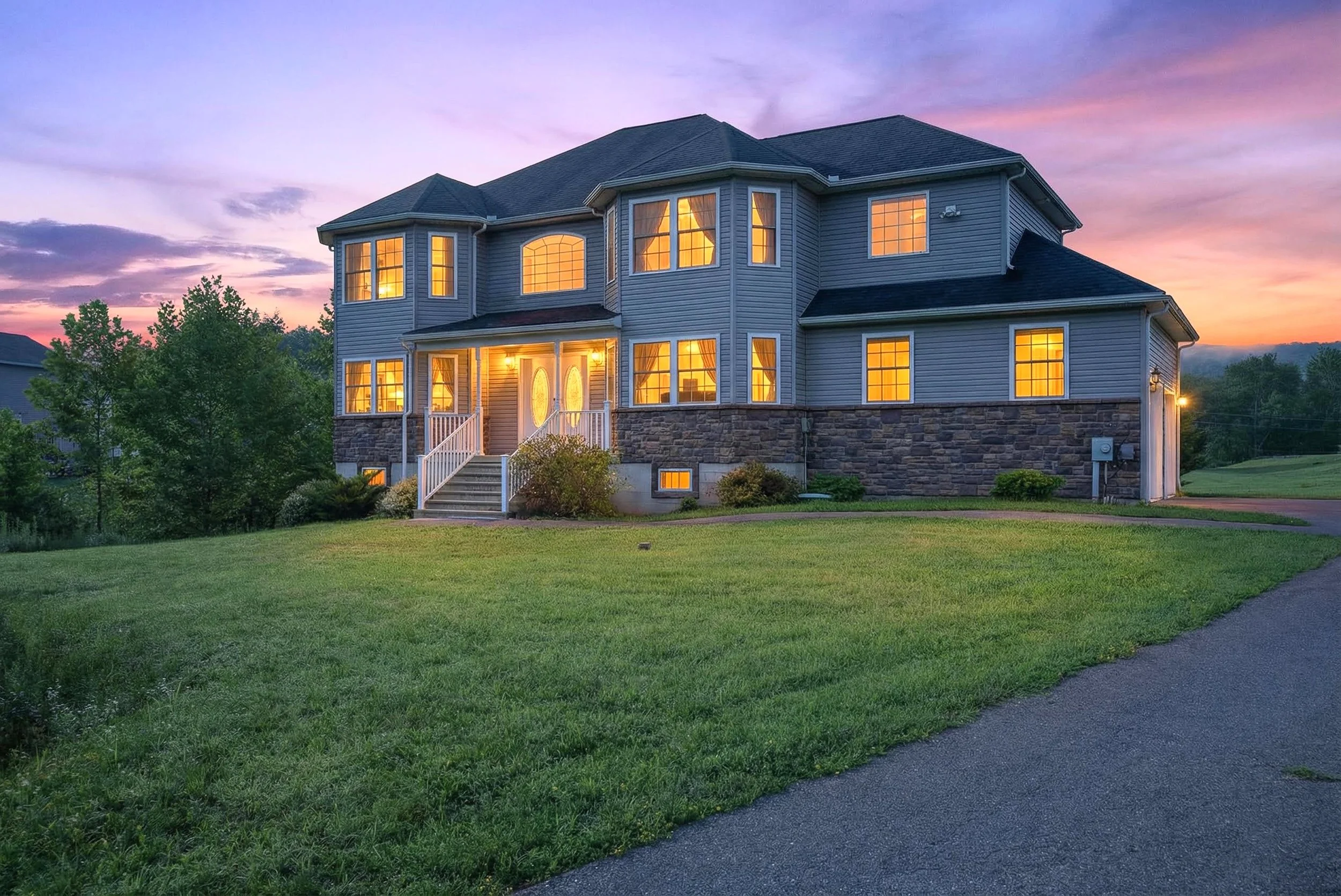 A large, multi-story house with blue siding and a stone foundation, illuminated from within, during sunset. The house has a front porch with stairs, multiple windows, and is surrounded by a well-maintained lawn.