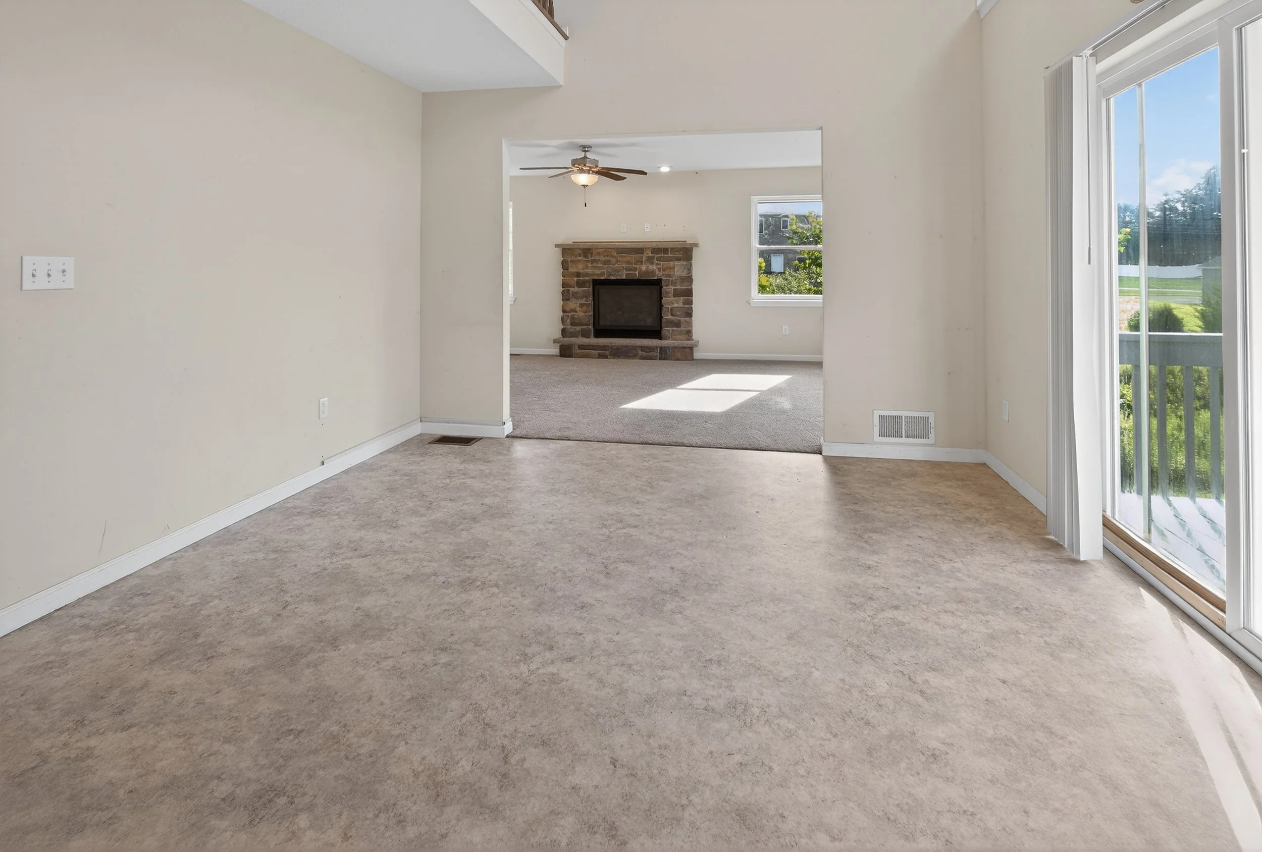 Empty living room with open view towards a fireplace and a window, sliding glass door leading outside, beige walls, carpeted and vinyl flooring.