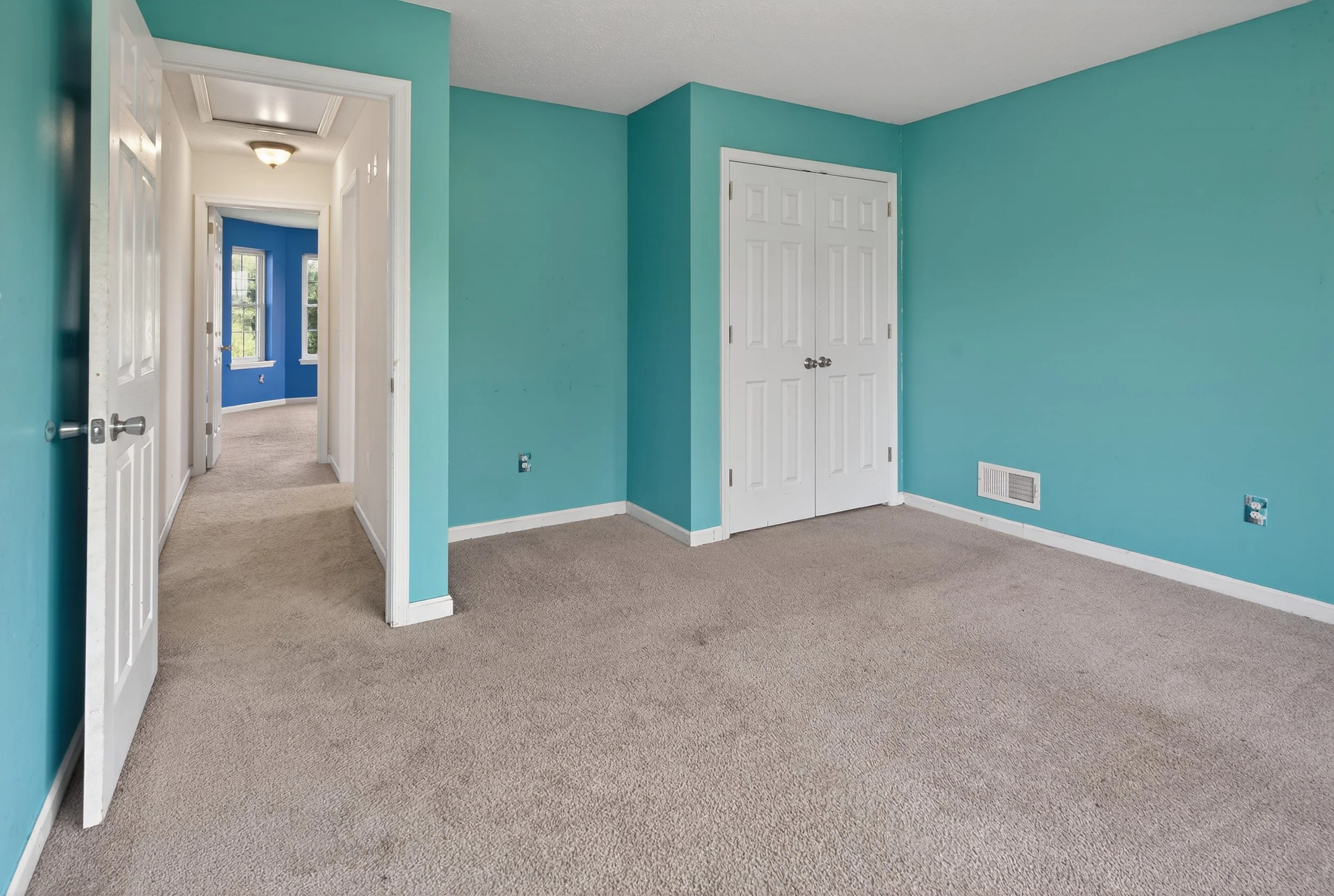 Empty room with light blue walls, white closet doors, beige carpet, and a white vent on the wall. An open door leads to a hallway with beige carpet and a room with blue walls and windows.