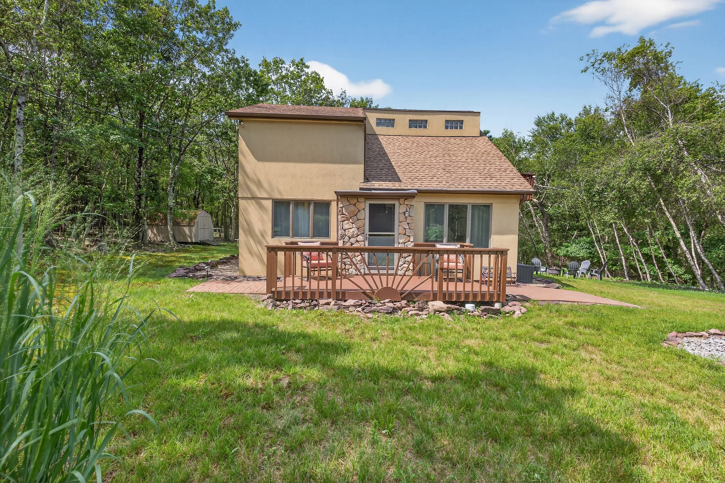 Front view of a house with a small wooden deck and grassy yard, surrounded by trees under a blue sky.