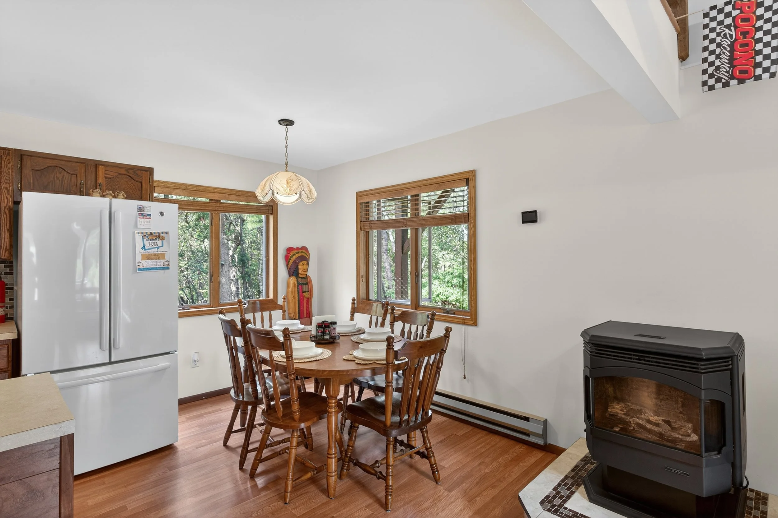 Dining room with a wooden table set with white plates and bowls, surrounded by wooden chairs, with windows showing a green outdoor view, a vintage hanging light fixture, a white refrigerator, and a black stove on a tiled hearth.