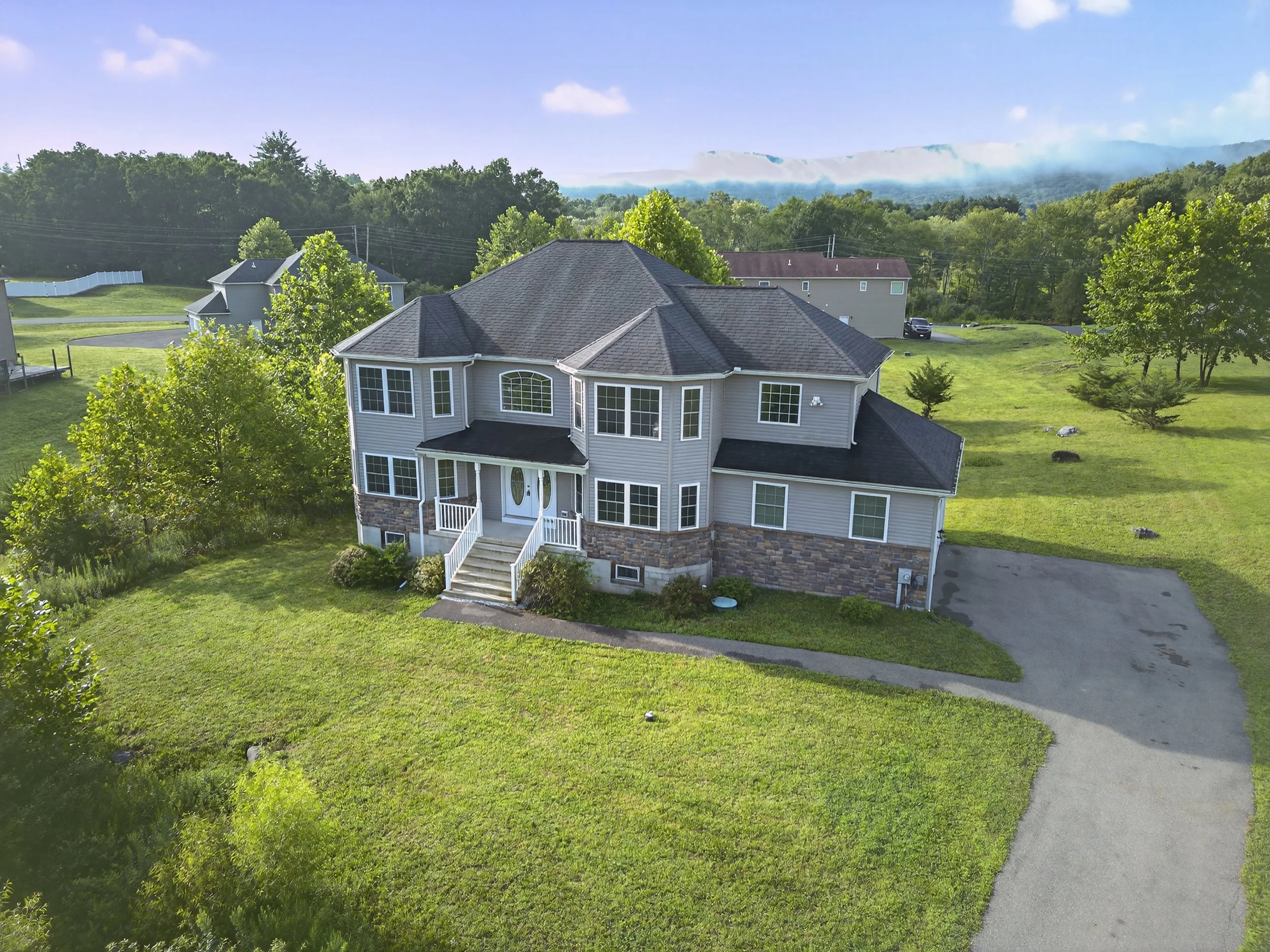 A large two-story house with a gray exterior and a black roof, with a front porch and white stairs leading to the door, surrounded by a green lawn and trees, with a driveway on the right and neighboring houses in the background.