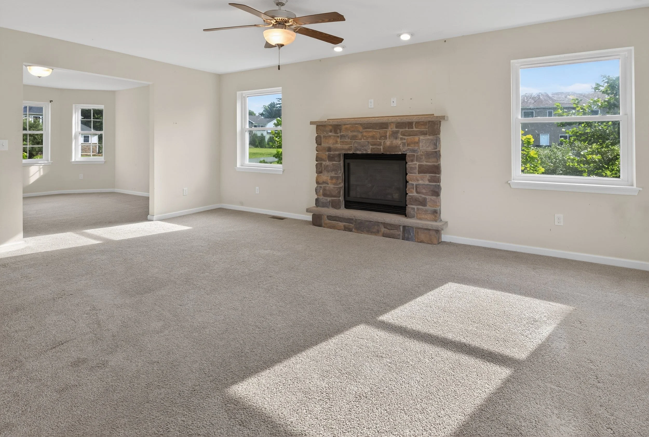 Empty living room with beige carpet, a stone fireplace, multiple windows showing green trees, and a ceiling fan.
