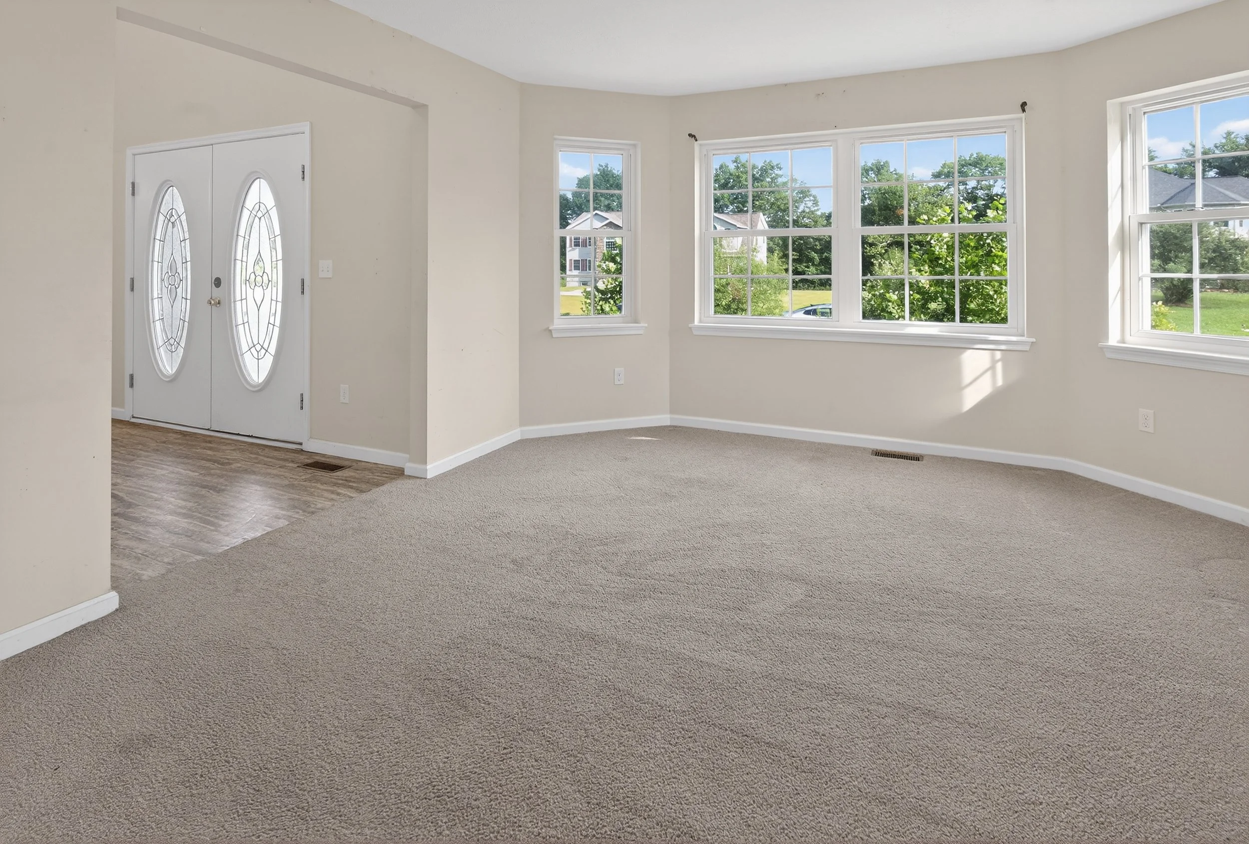Empty living room with large bay windows, front door with glass panels, beige carpet, and cream-colored walls.