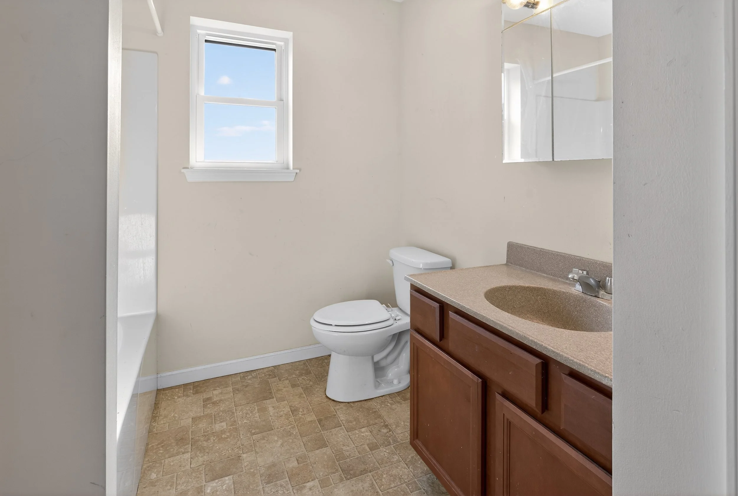A small bathroom with a window, a toilet, a brown vanity with a sink, a mirror, and a shower area, with beige tiled flooring.