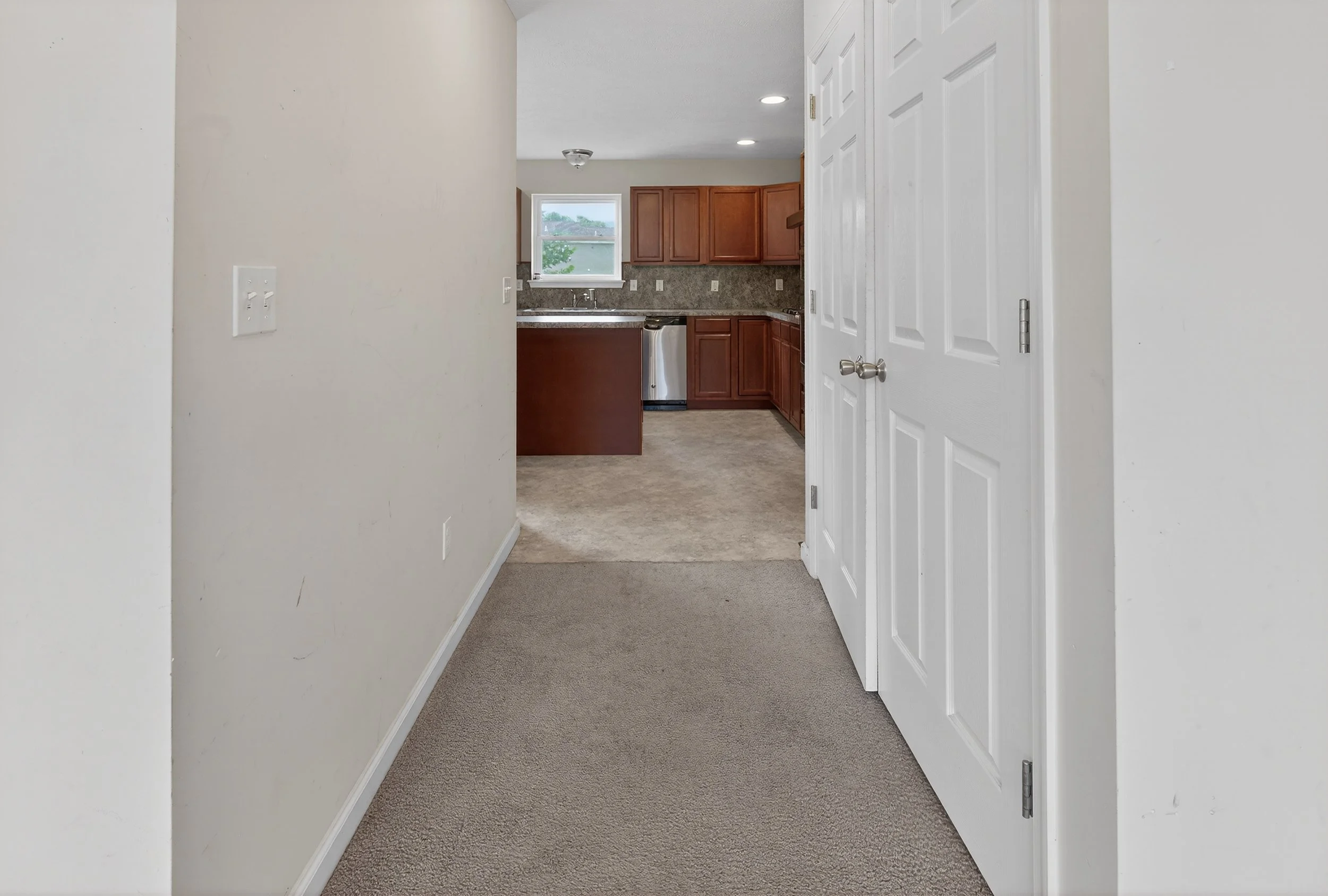 A view of a hallway leading into a kitchen with brown cabinets and a window, with white double closet doors on the right.