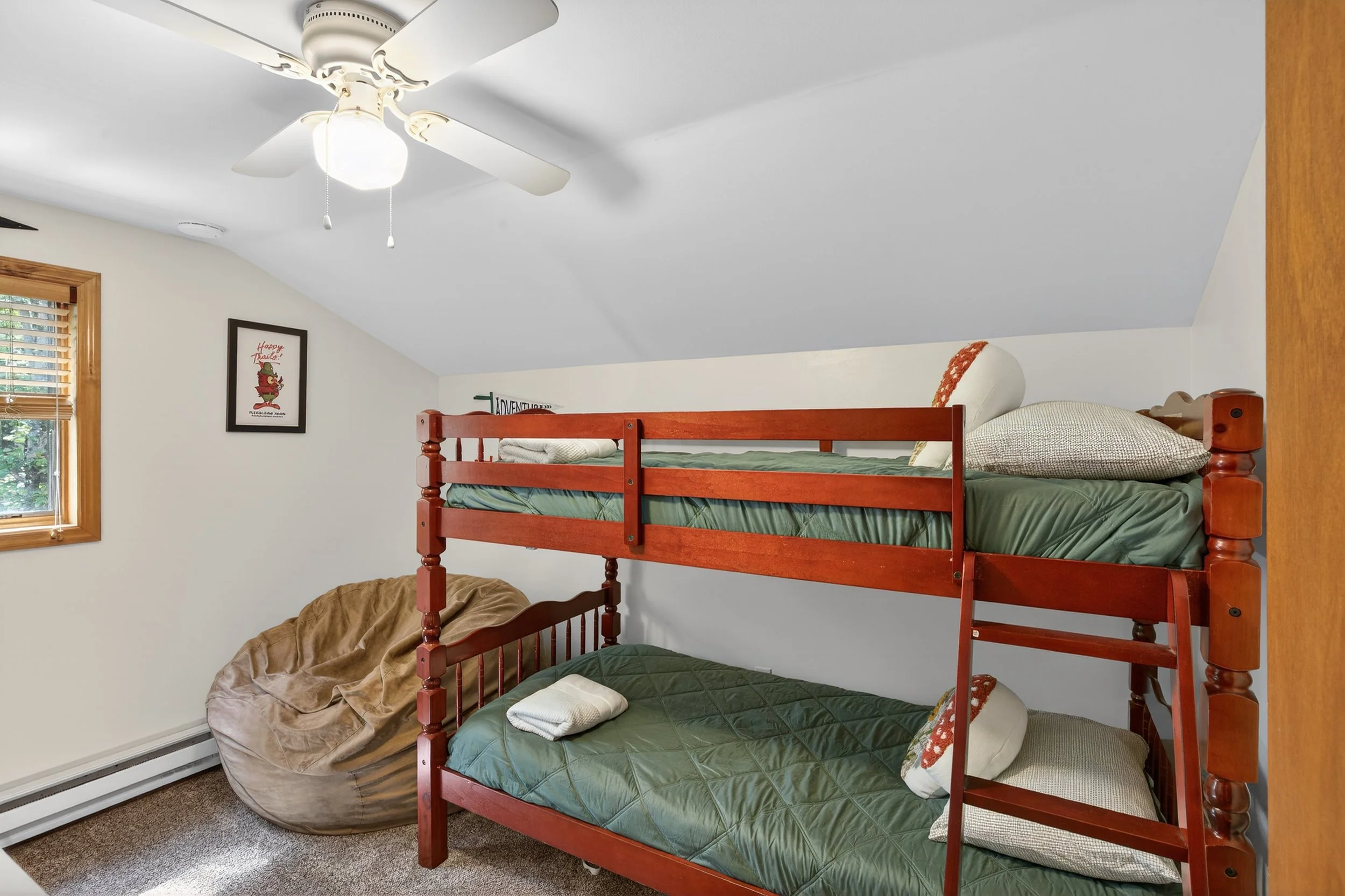 Room with white sloped ceiling, ceiling fan with light, small window with wooden blinds, framed picture on the wall, and bunk beds with green mattresses, pillows, and a foldable ladder. A beige bean bag chair is on the carpeted floor.