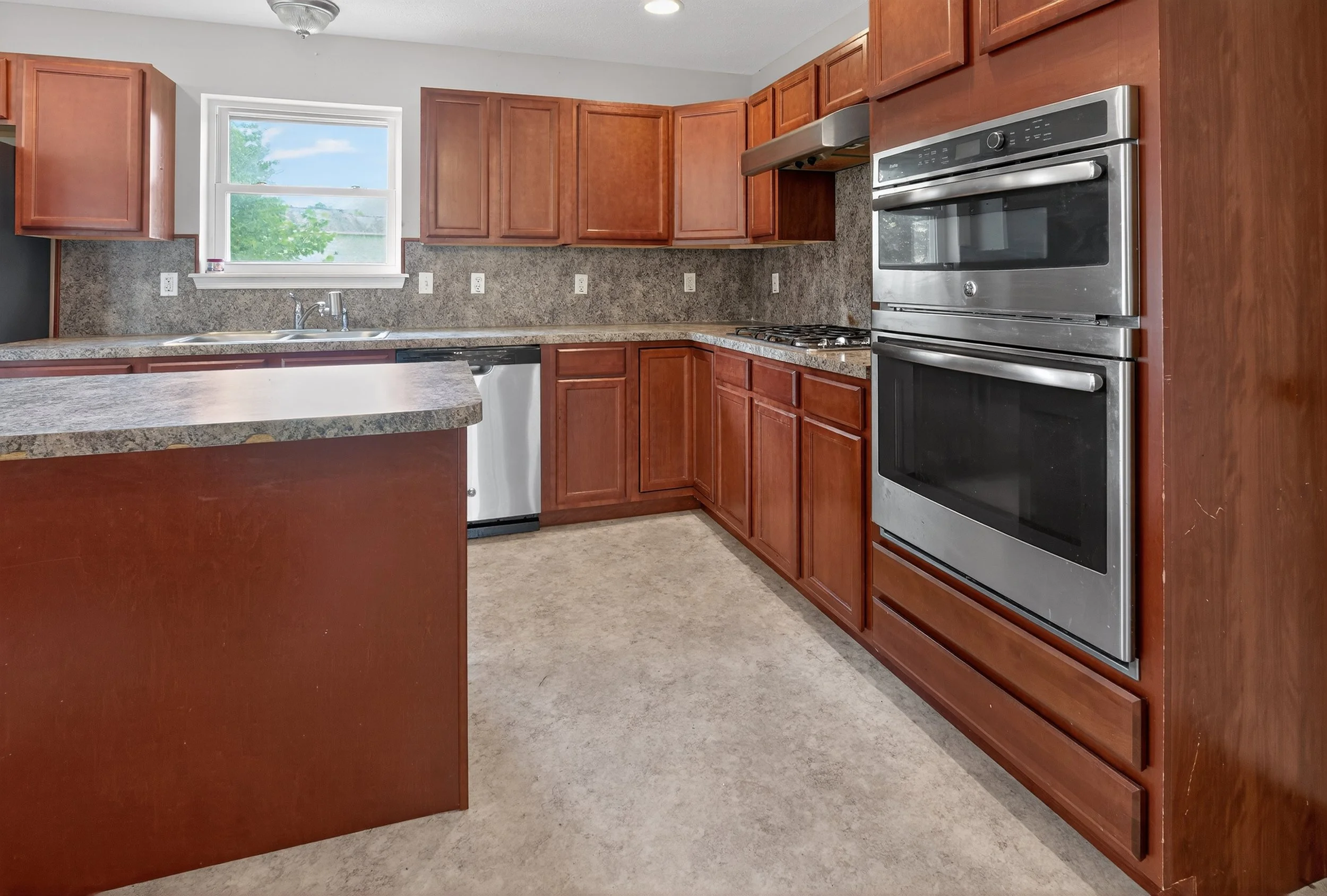 Kitchen with brown wooden cabinets, granite countertops, a window above the sink, stainless steel double oven, and a dishwasher.