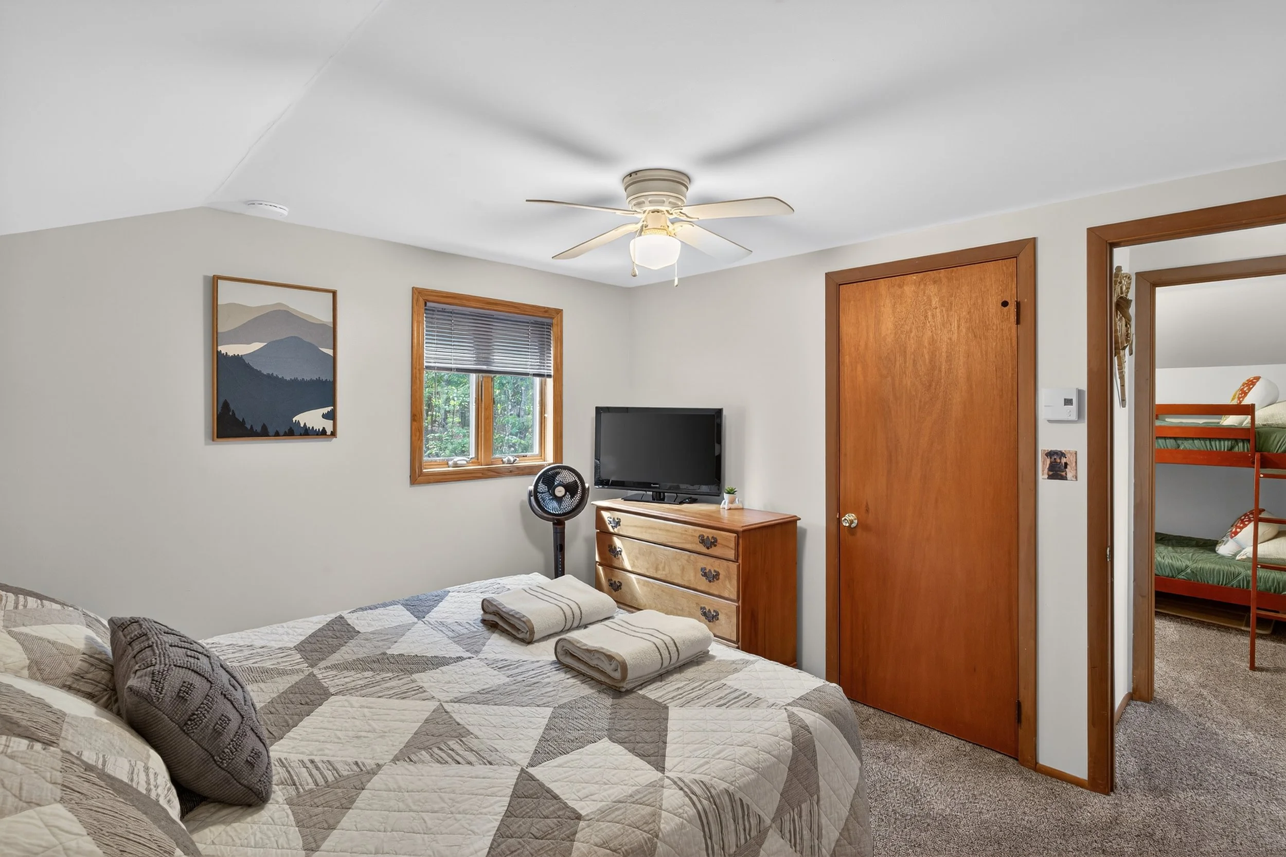 Bedroom with beige walls, wooden trim, bed with patterned quilt and pillows, three towels on bed, window with blinds, small television on wooden dresser, ceiling fan