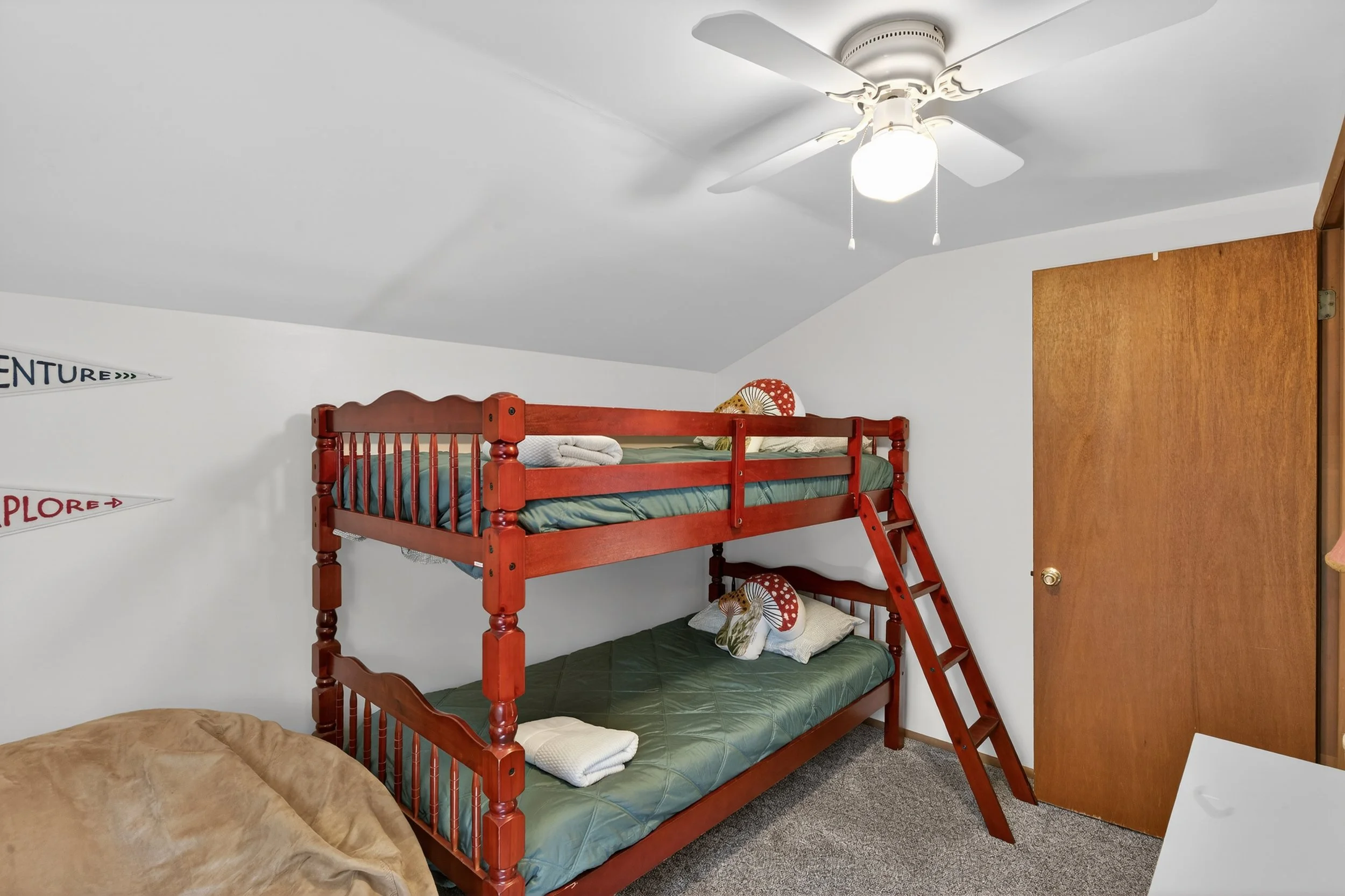 Bedroom with a wooden bunk bed, a ceiling fan with light, wooden closet door, and partial view of a beige chair or bed in the foreground.