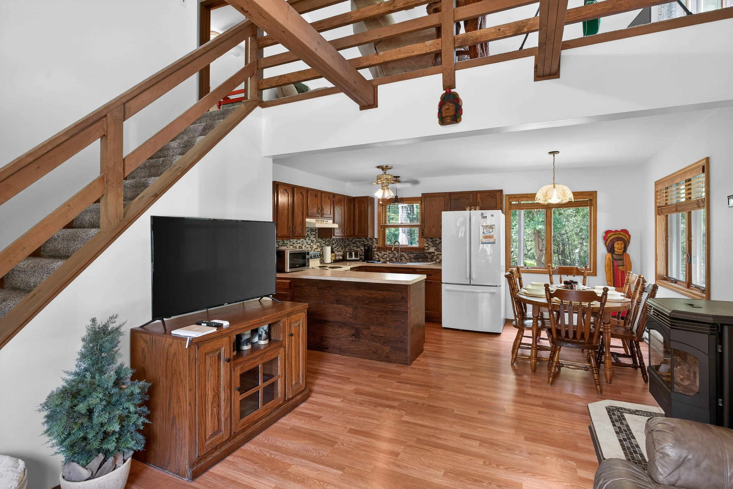 Living area with a wooden TV stand, a flat-screen TV, a small potted plant, a wood staircase leading upstairs, a kitchen with wooden cabinets, a white refrigerator, a dining table with wooden chairs, a window with blinds, and a stove.