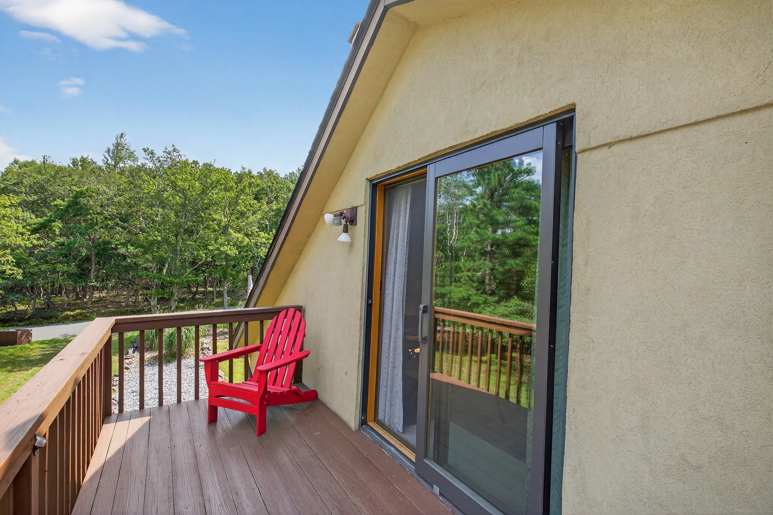 A small balcony with a red Adirondack chair overlooking a lush green yard and trees, with a yellow exterior wall and a sliding glass door.