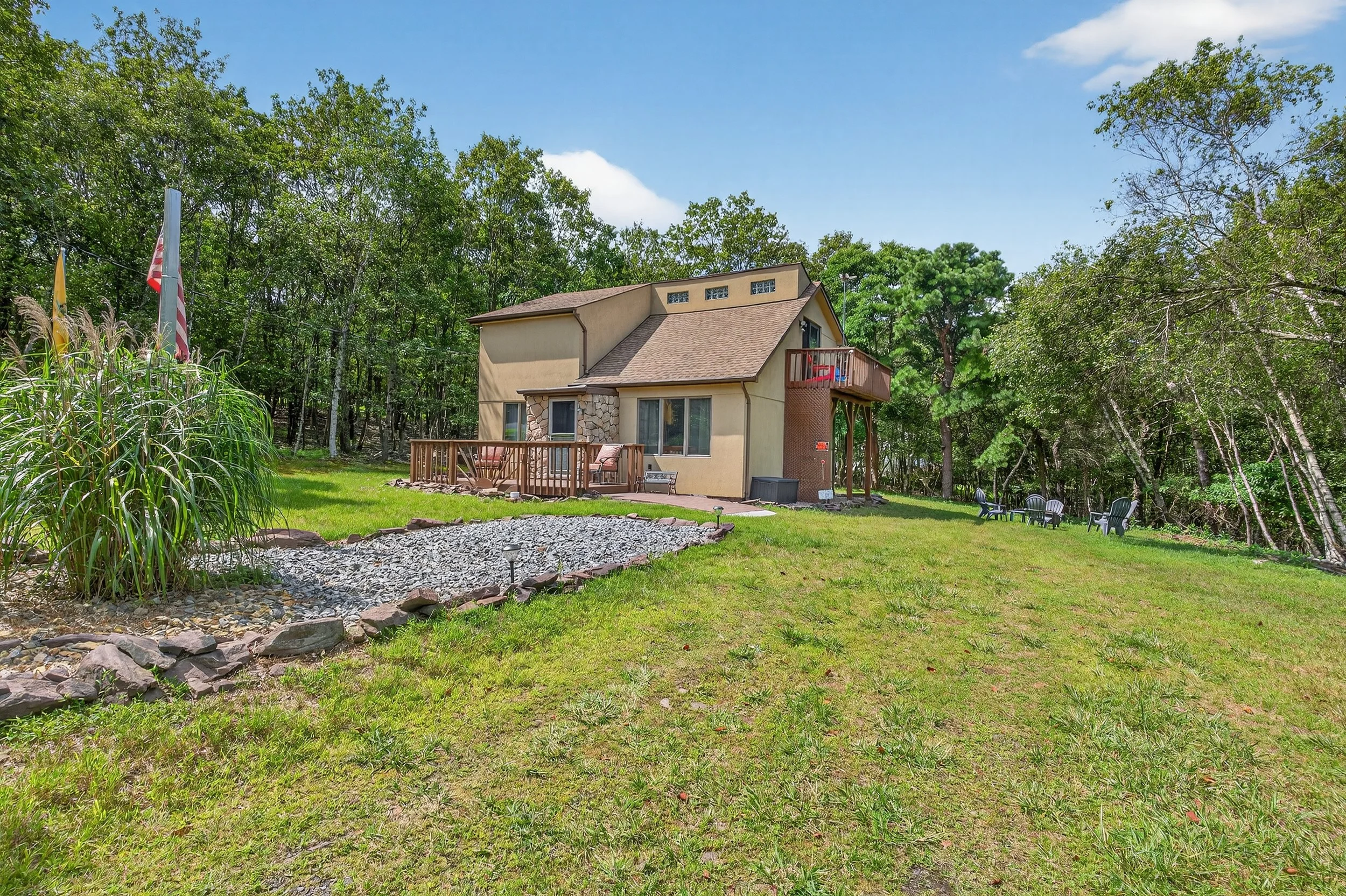 Backyard of a house with a grassy lawn, a small deck with outdoor furniture, surrounded by trees, with a flag and flags on poles to the left, and a sitting area with chairs to the right.