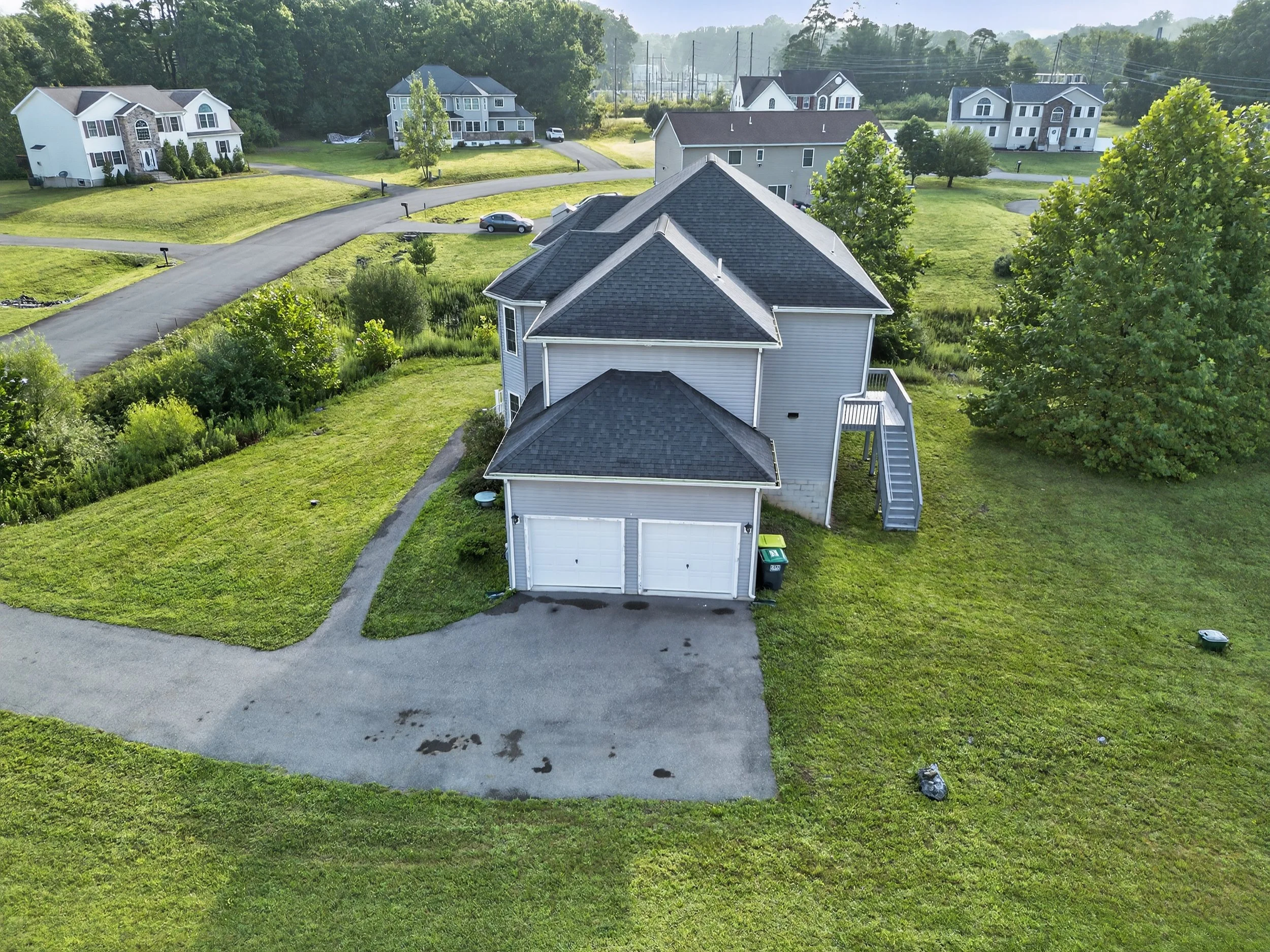 Aerial view of a suburban neighborhood featuring a detached two-car garage attached to a house with a dark shingle roof, surrounded by green lawns and trees, with multiple other houses and streets in the background.