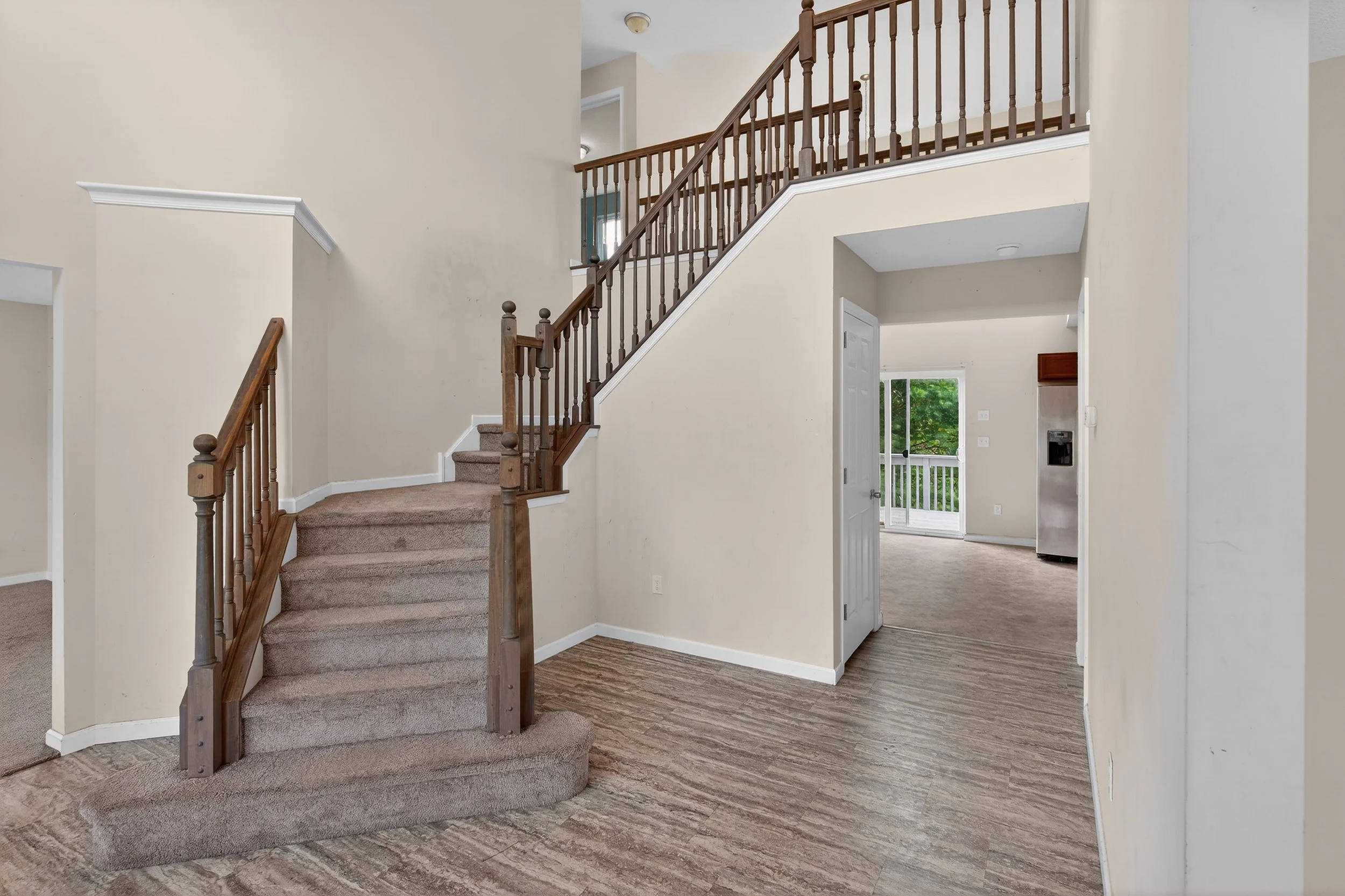 Interior view of a house showing a staircase with wooden railing, carpeted stairs, brown flooring, and a small kitchen area with a stainless steel refrigerator and sliding door leading outside