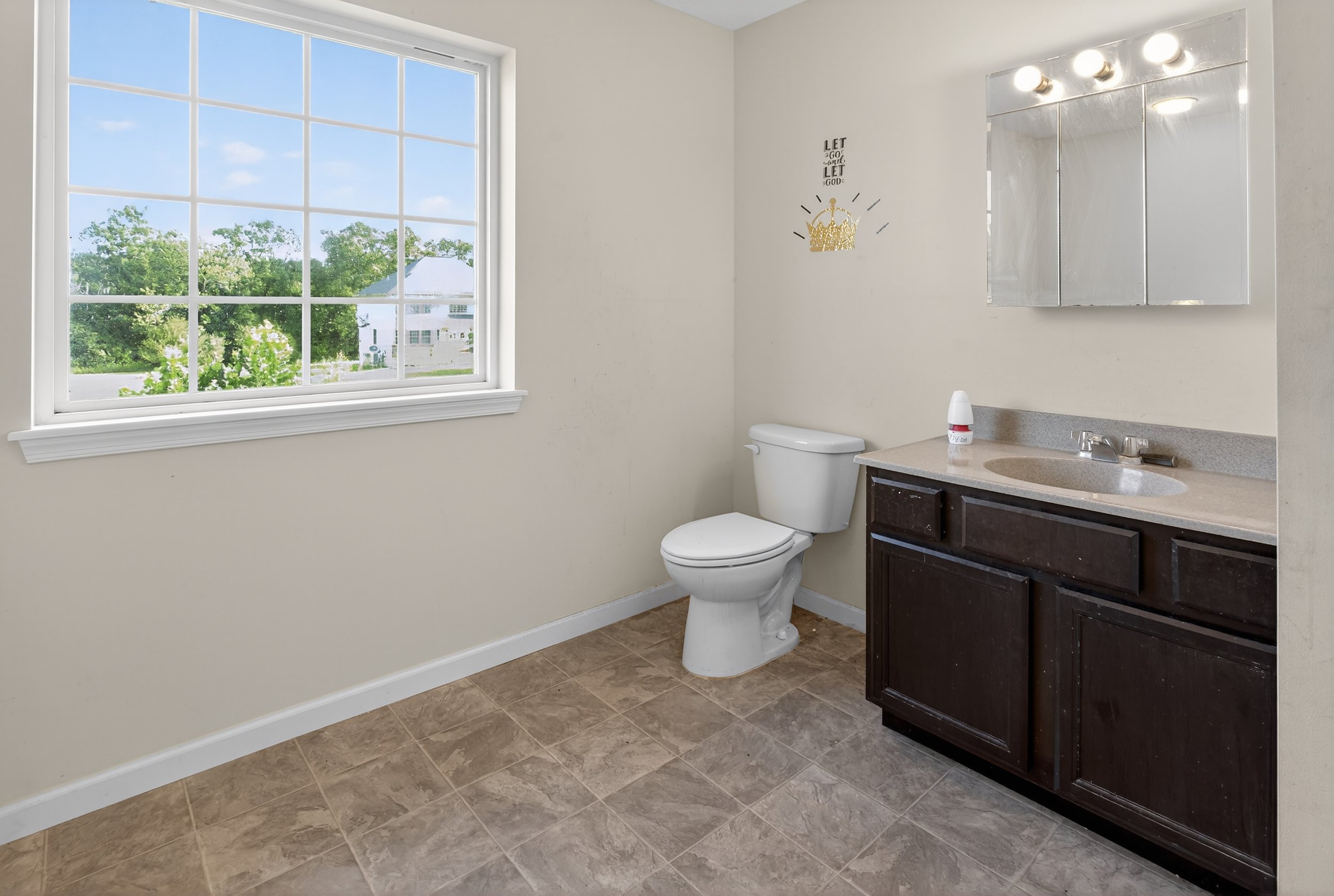 A bathroom with a window, toilet, and dark wooden vanity with a sink, mirror, and bathroom supplies, illuminated by three overhead lights.
