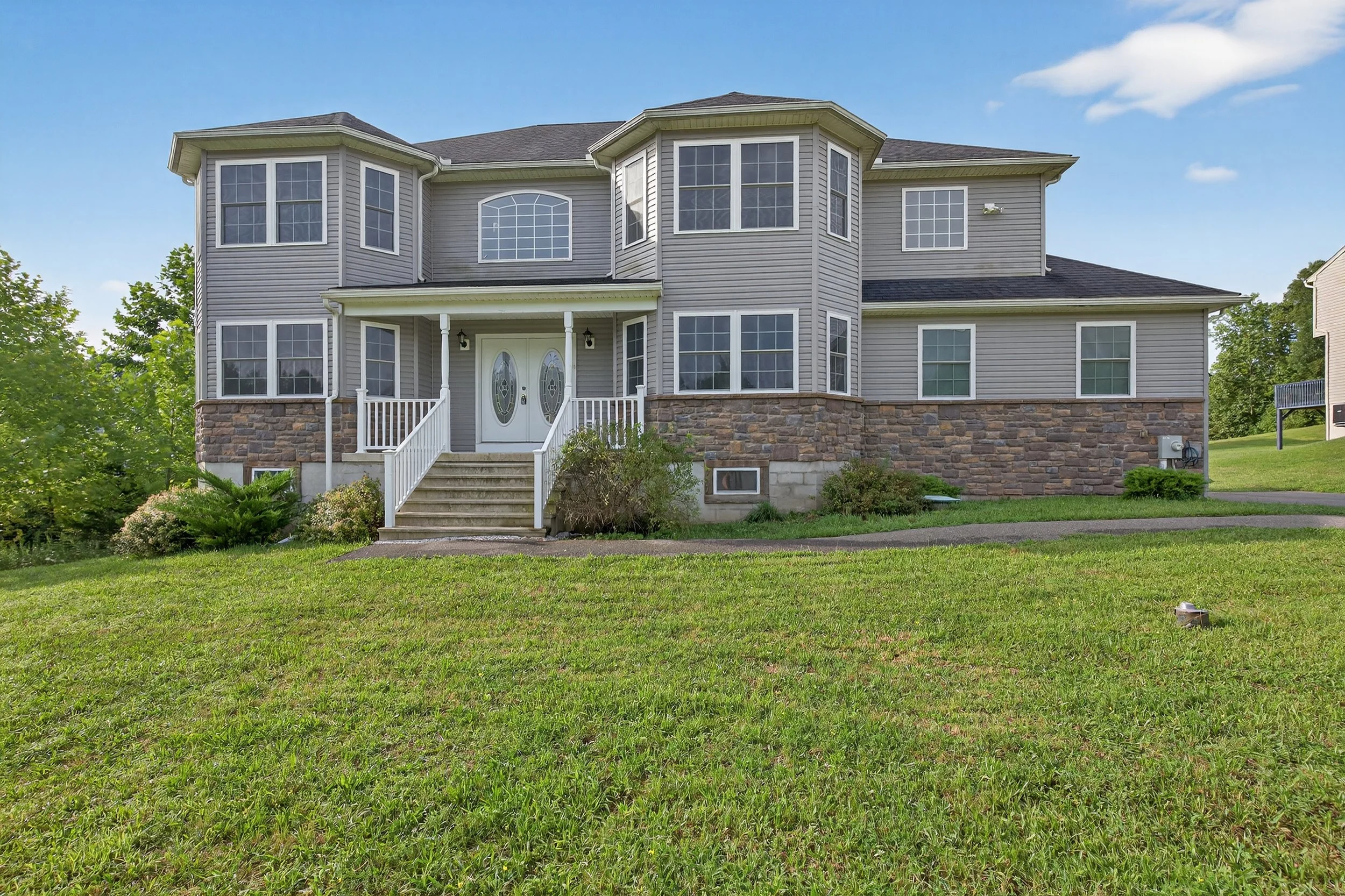 A large two-story house with gray siding, white trim, and a stone foundation, featuring several bay windows, a front porch with stairs, and a well-maintained lawn.