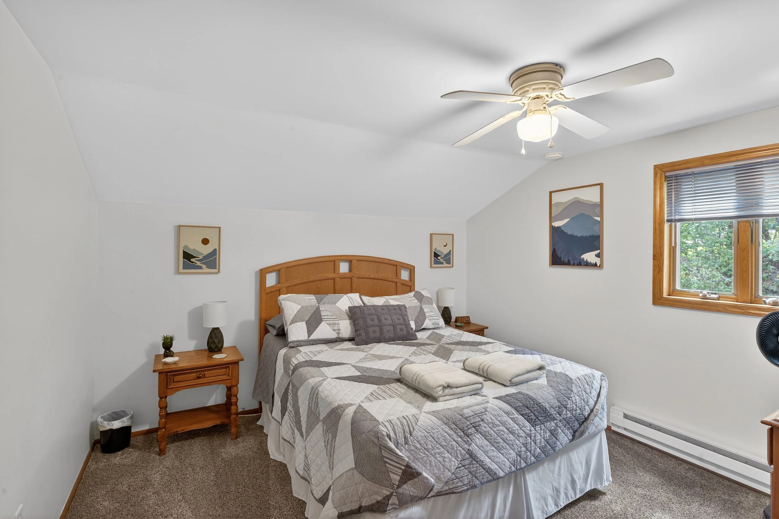 A bedroom with a wooden bed frame, a patterned quilt, and folded towels on the bed. There are two wooden nightstands with lamps, a window with wooden trim and a gray blind, a wall art print, and a ceiling fan.